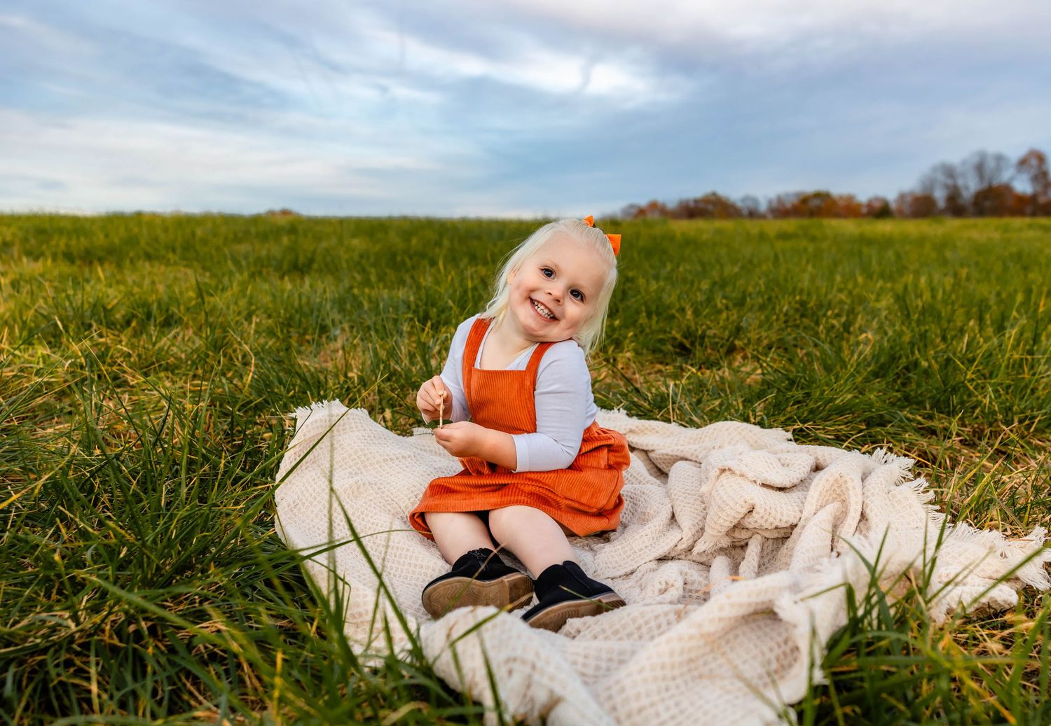 A toddler in an orange jumper dress sits on a white blanket in a grassy field on a sunny day.
