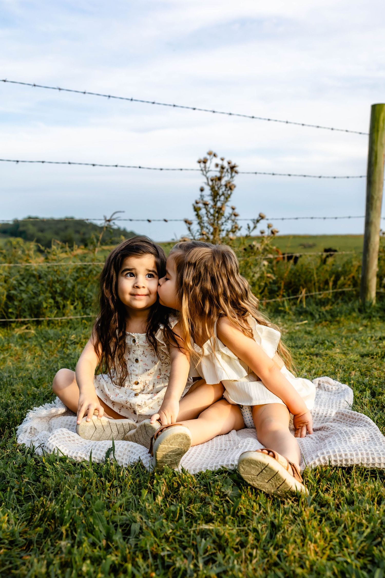Children in summer dresses sharing secrets while sitting on blanket in rustic outdoor setting.