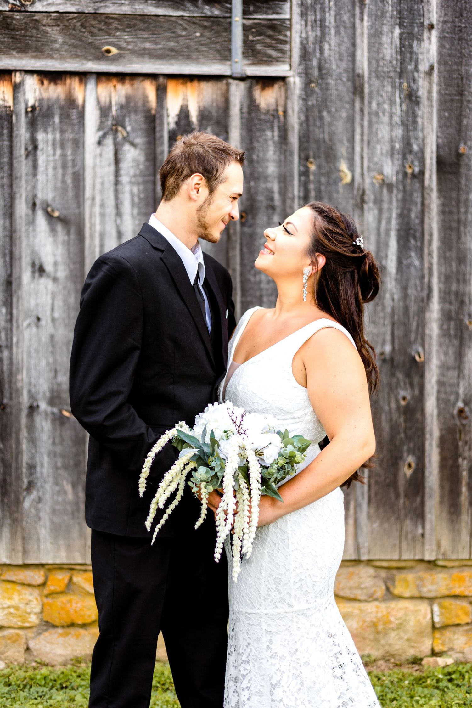A romantic wedding photo of a bride and groom sharing a moment against a rustic wooden barn wall.