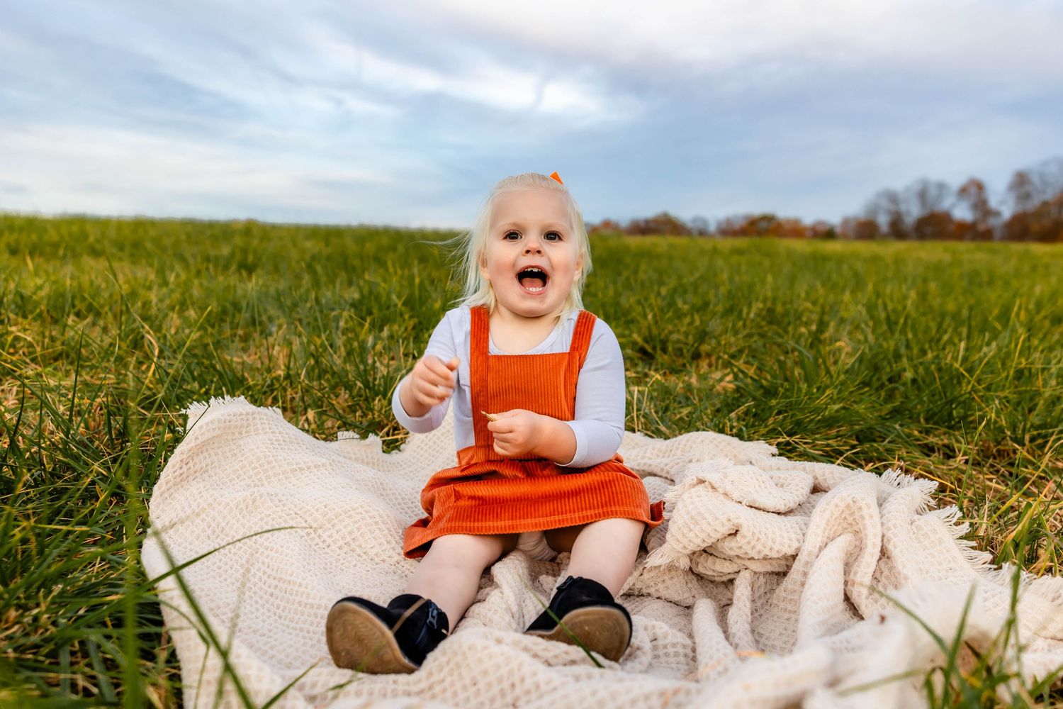 Excited toddler in orange pinafore dress sitting on white blanket in grassy field.