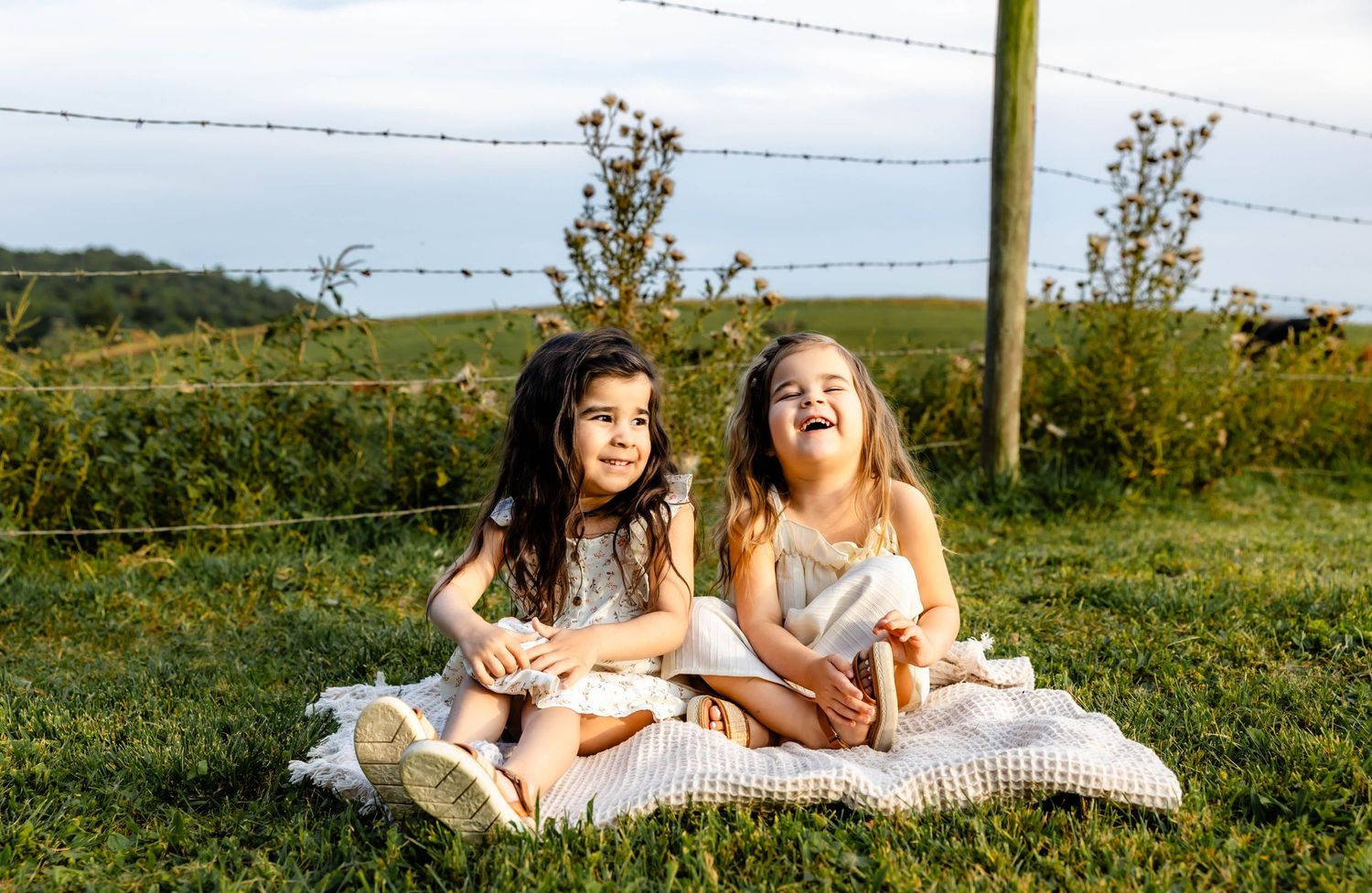 Two children sitting together on white blanket in scenic countryside setting during golden hour.