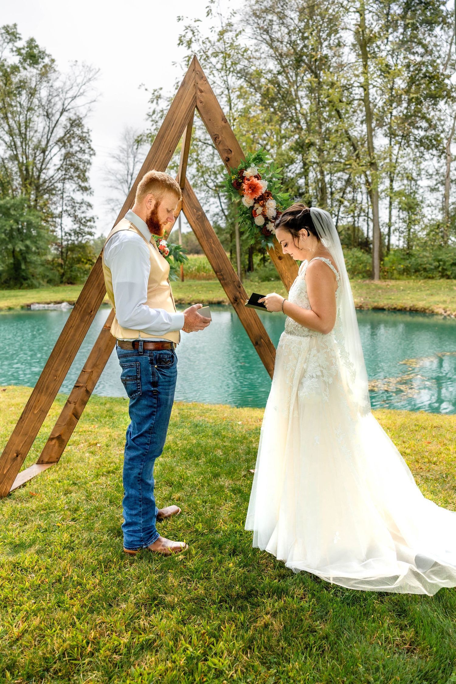 Wedding ceremony moment captured under a triangular wooden arch by a peaceful pond.