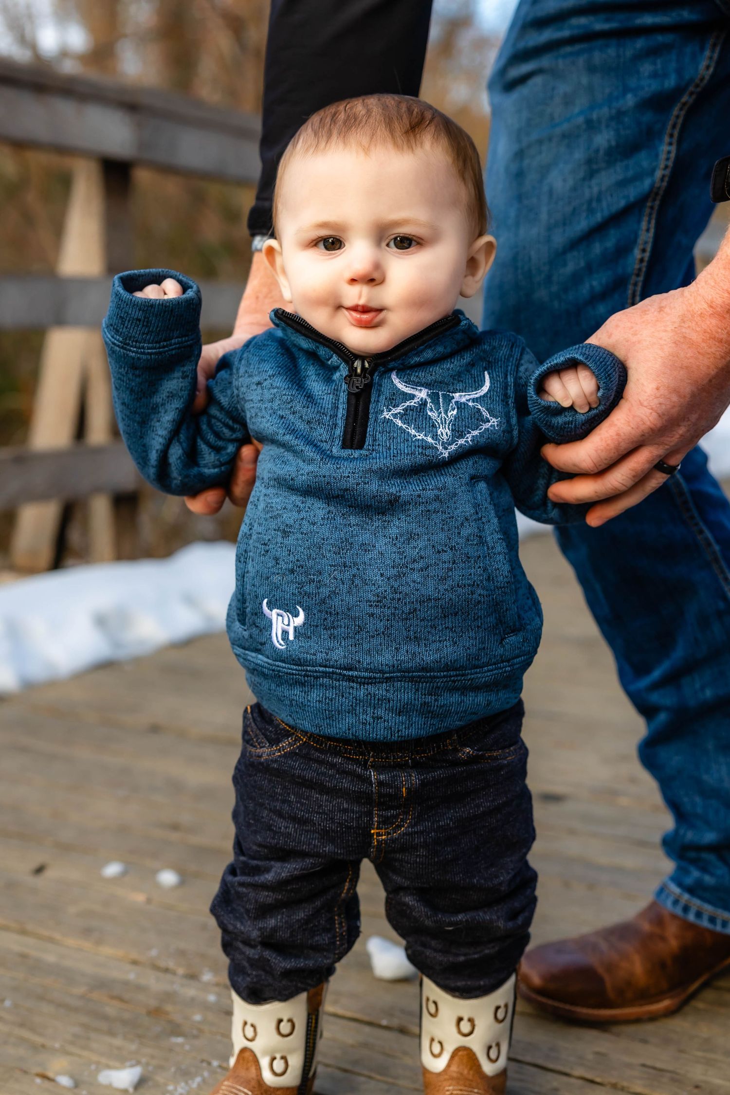 Small child wearing blue half-zip sweater and boots standing on wooden bridge.