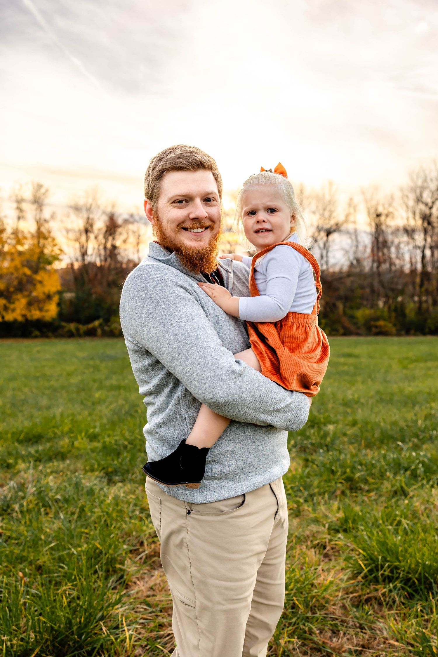 A tender outdoor portrait showing an embrace between a parent and child in a gray sweater and orange jumper at sunset.