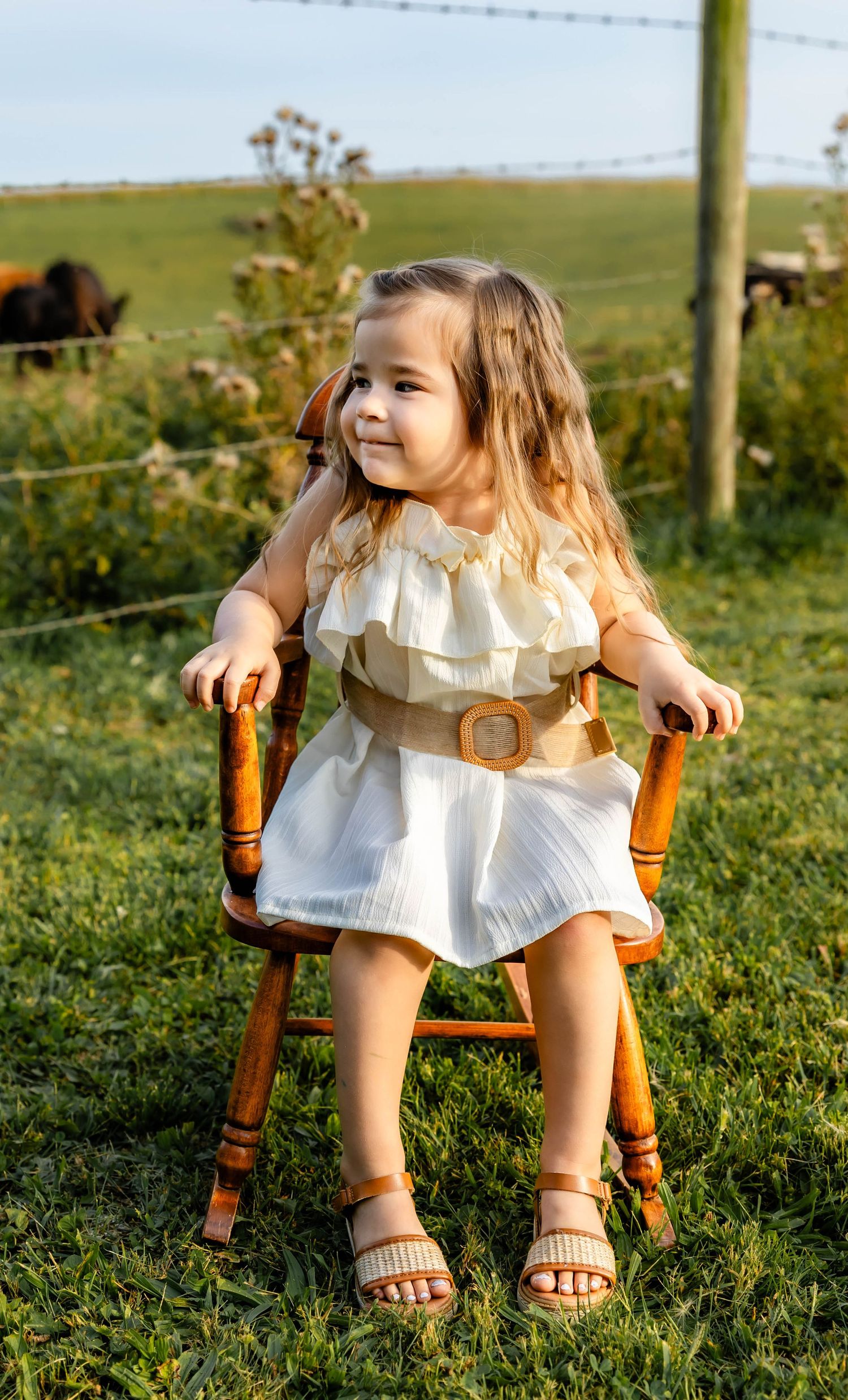Summer portrait in a white dress with belt detail, posed on wooden chair in farm setting.