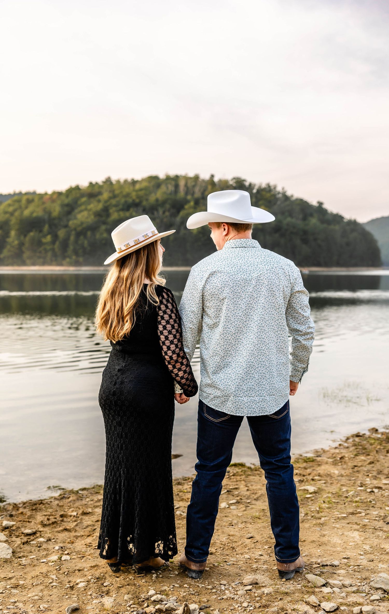 Couple in western wear stand hand in hand looking out over a serene mountain lake.