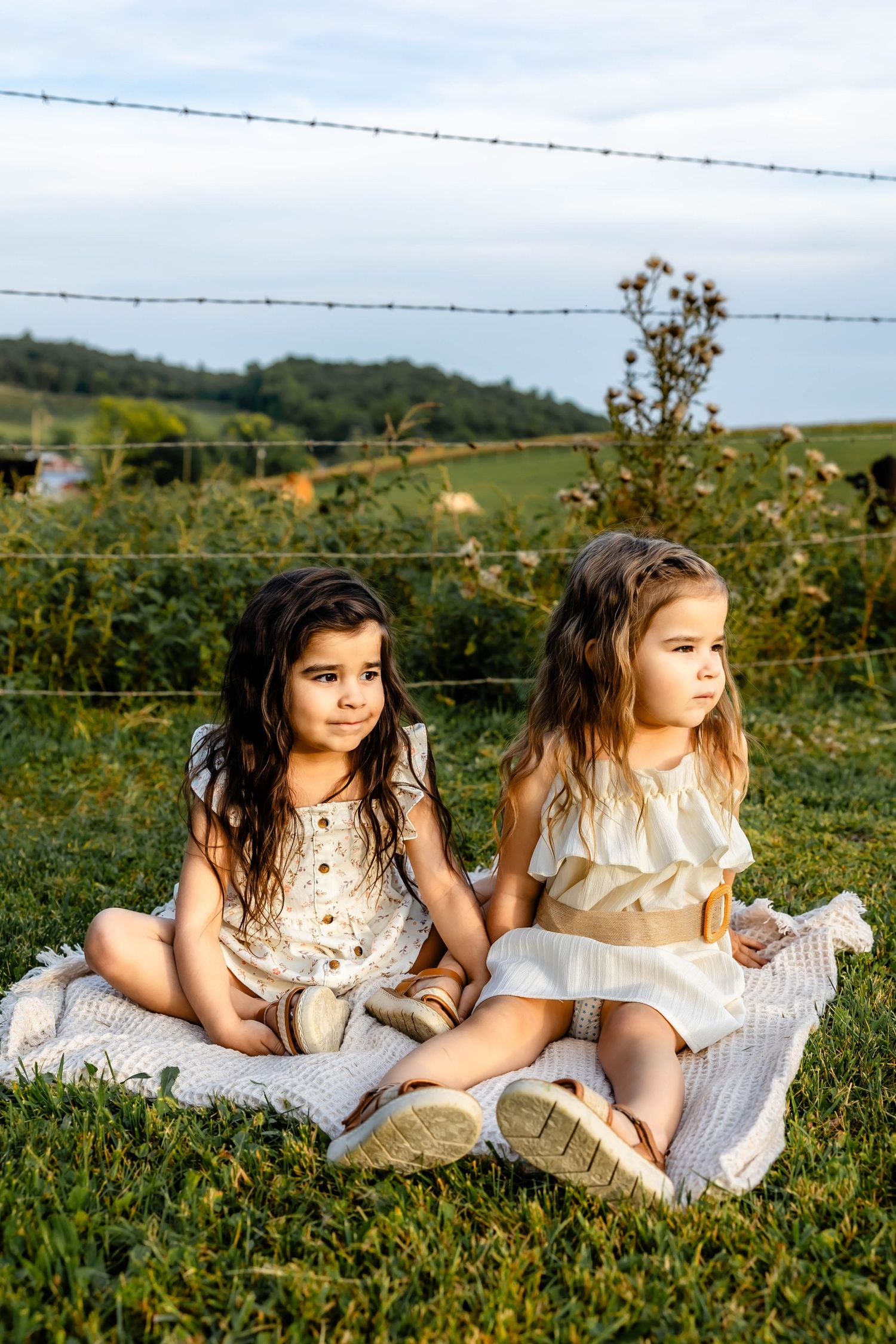 Children in summer outfits sitting side by side on white blanket against pastoral landscape.