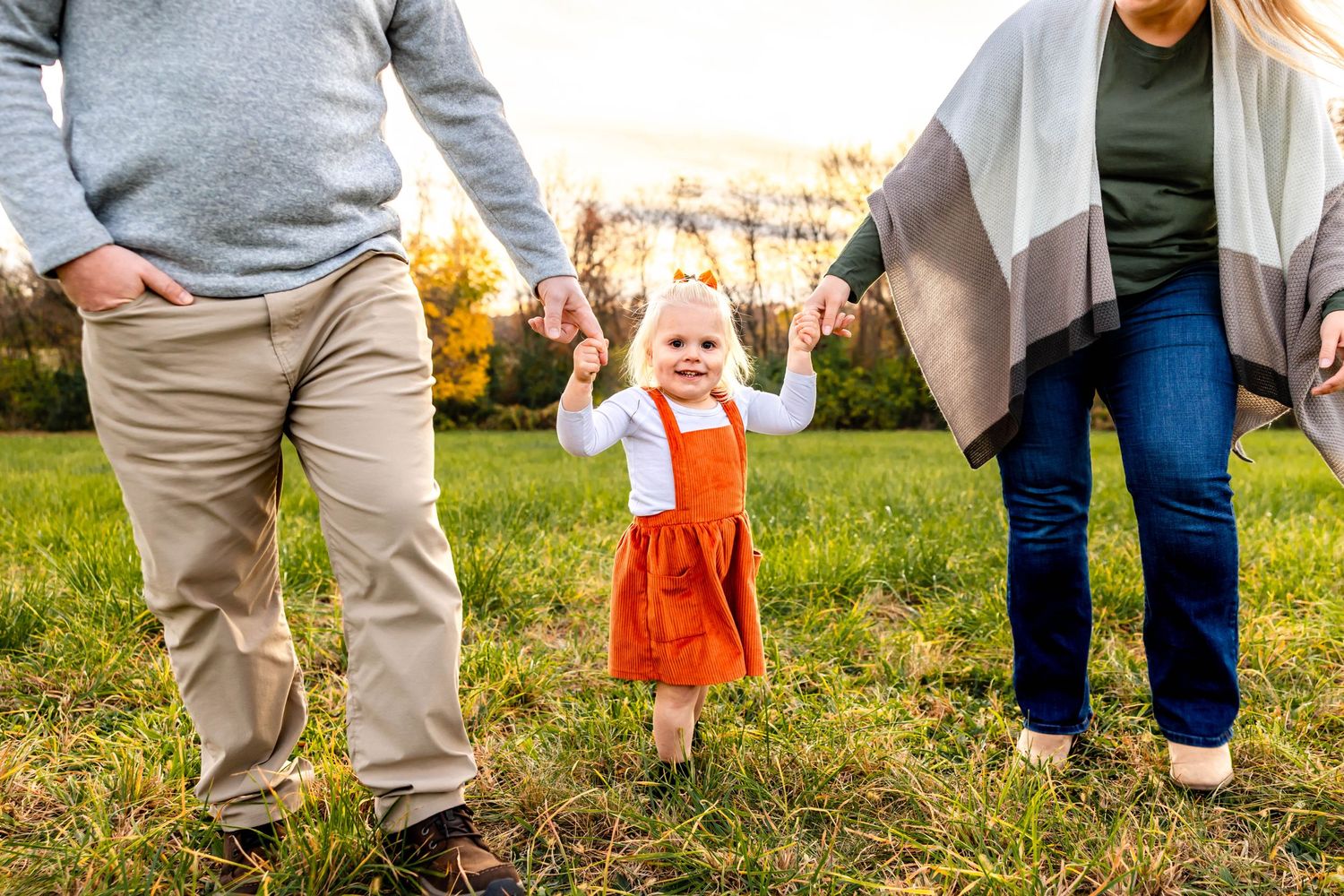 Family holds hands with child in orange jumper while walking through autumn field.