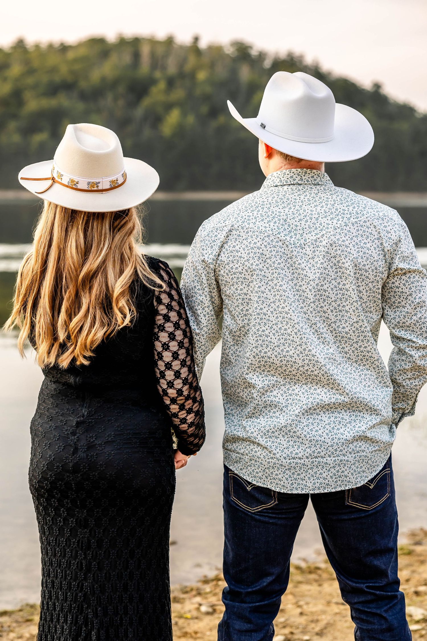 Back view of couple in western wear and cowboy hats standing by a lake.