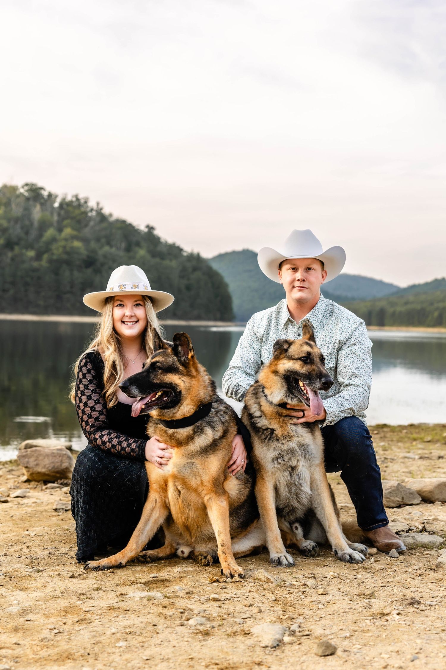 Portrait of a couple in western wear posing with their German Shepherd dogs by a serene lake.