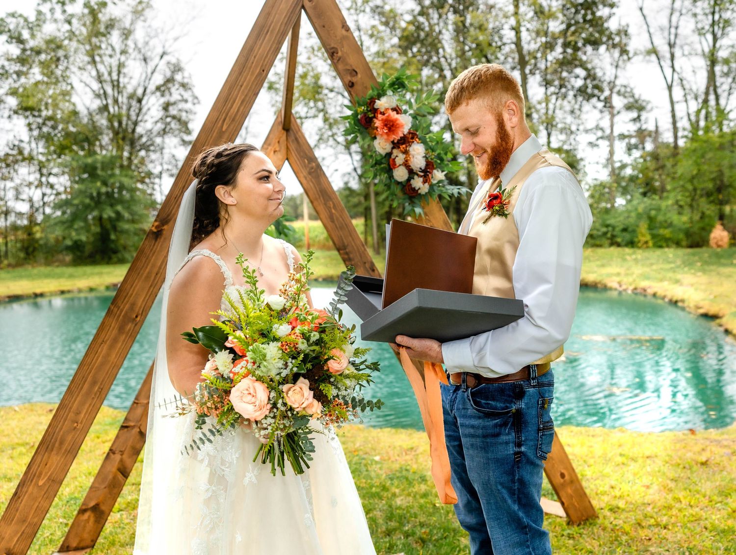 Wedding ceremony with wooden triangle arch and colorful floral arrangements beside a blue pond.