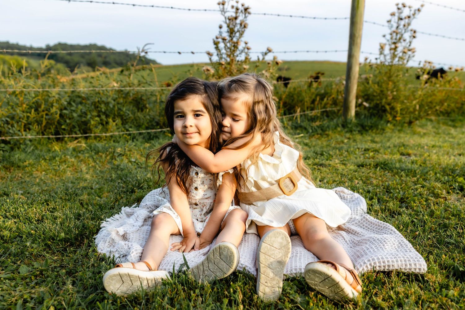 Two children in white dresses sharing a playful moment while sitting on blanket in grassy field.
