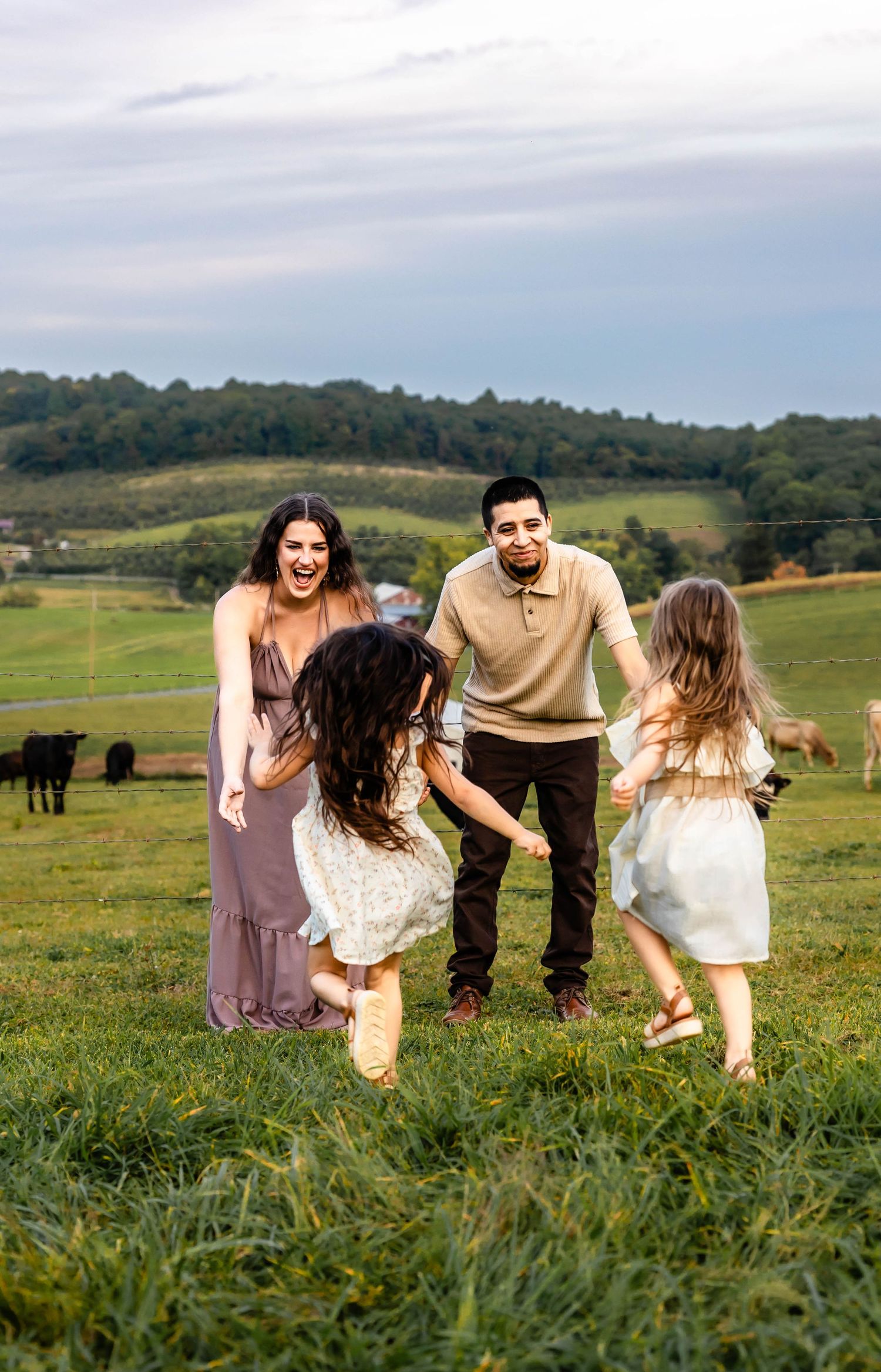 A family plays together in a grassy meadow with rolling hills and cattle in the distance.