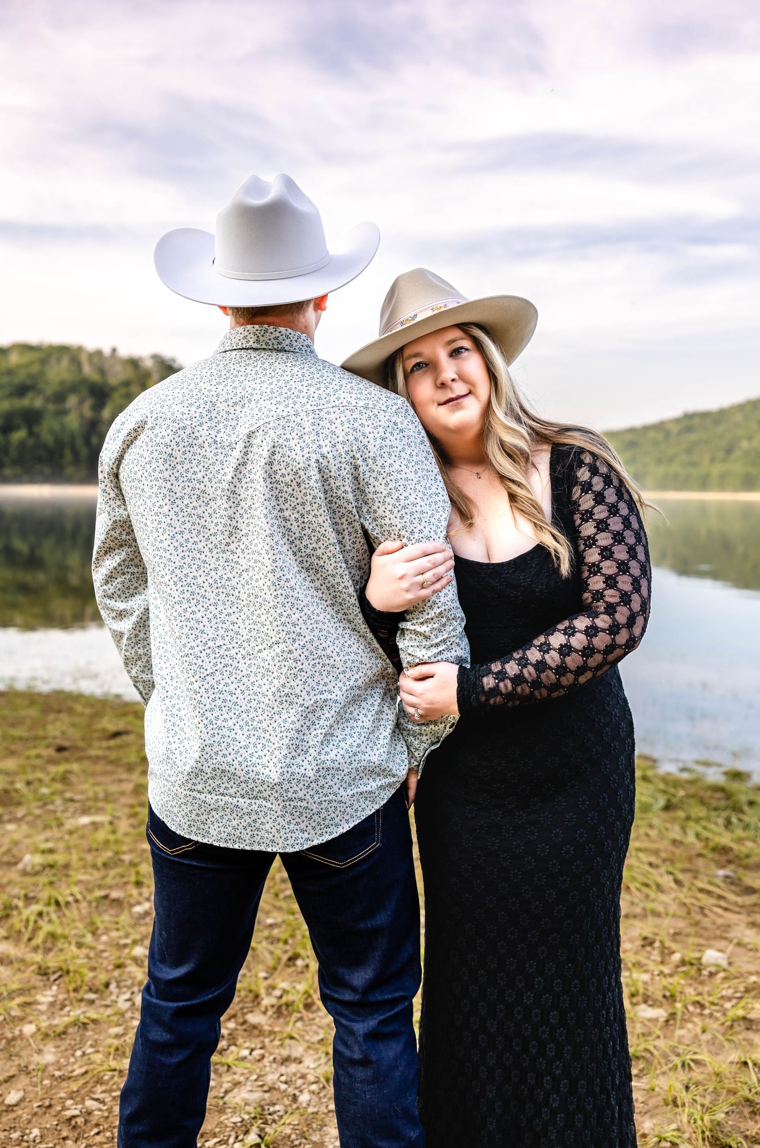 A couple in western attire embraces by a serene lake with mountains in the background.
