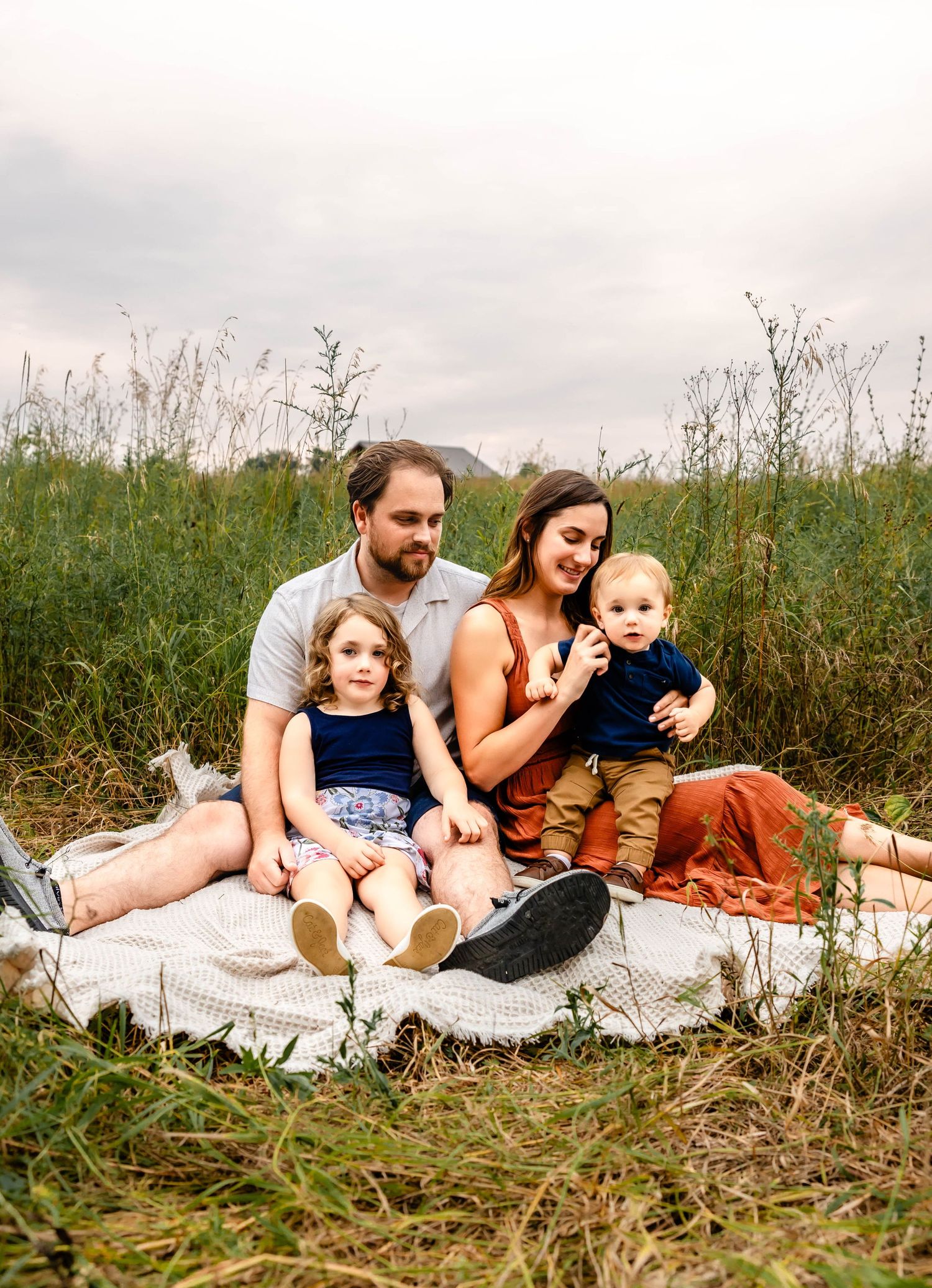 A family of four sits together on a blanket in a meadow during a casual outdoor photo session.