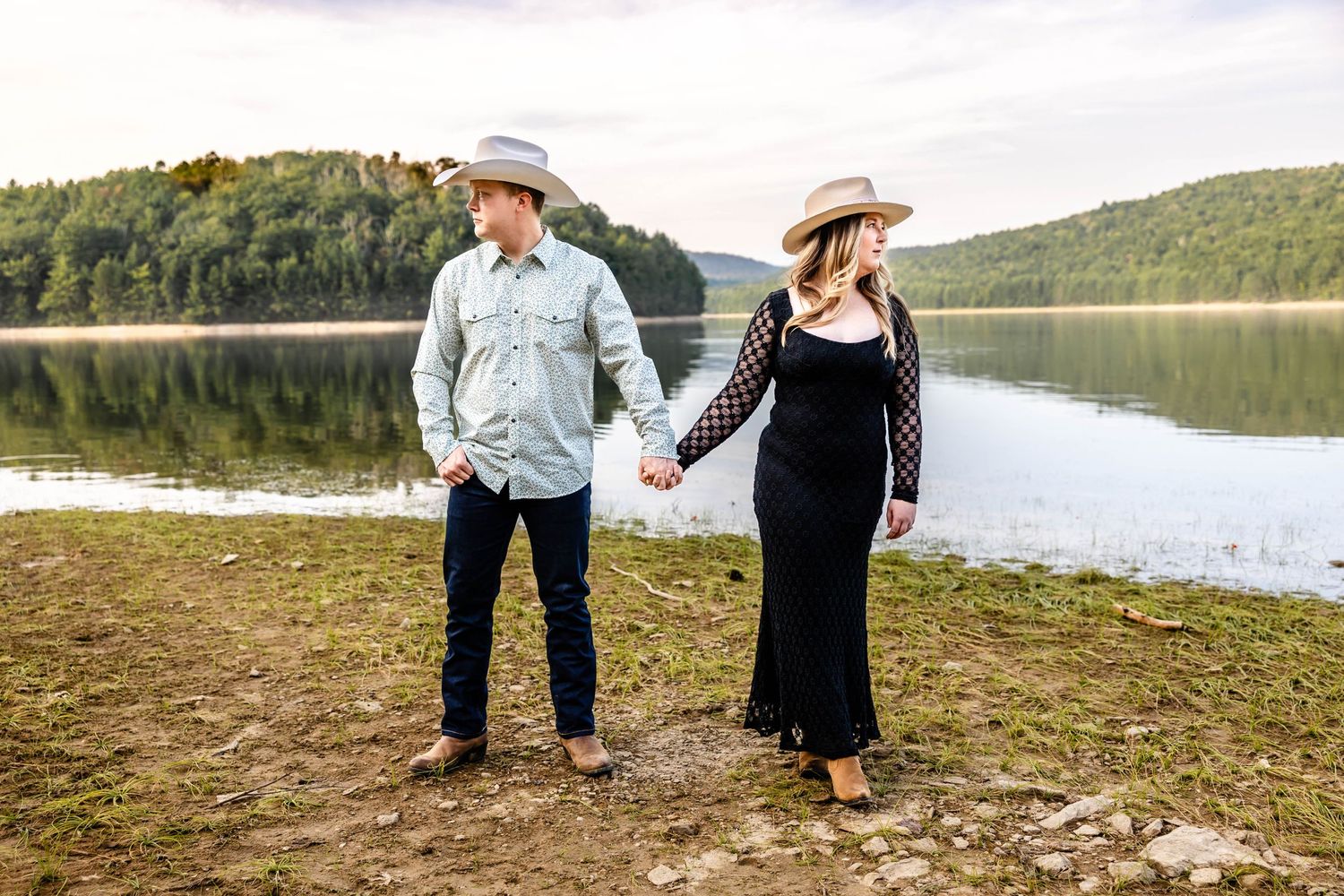 Couple in western wear holding hands while walking along a lakeside trail.