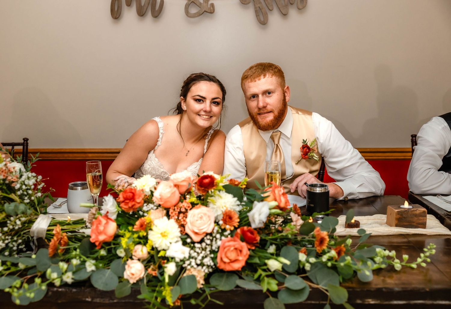 Wedding couple sits at rustic head table adorned with lush floral centerpiece arrangements.