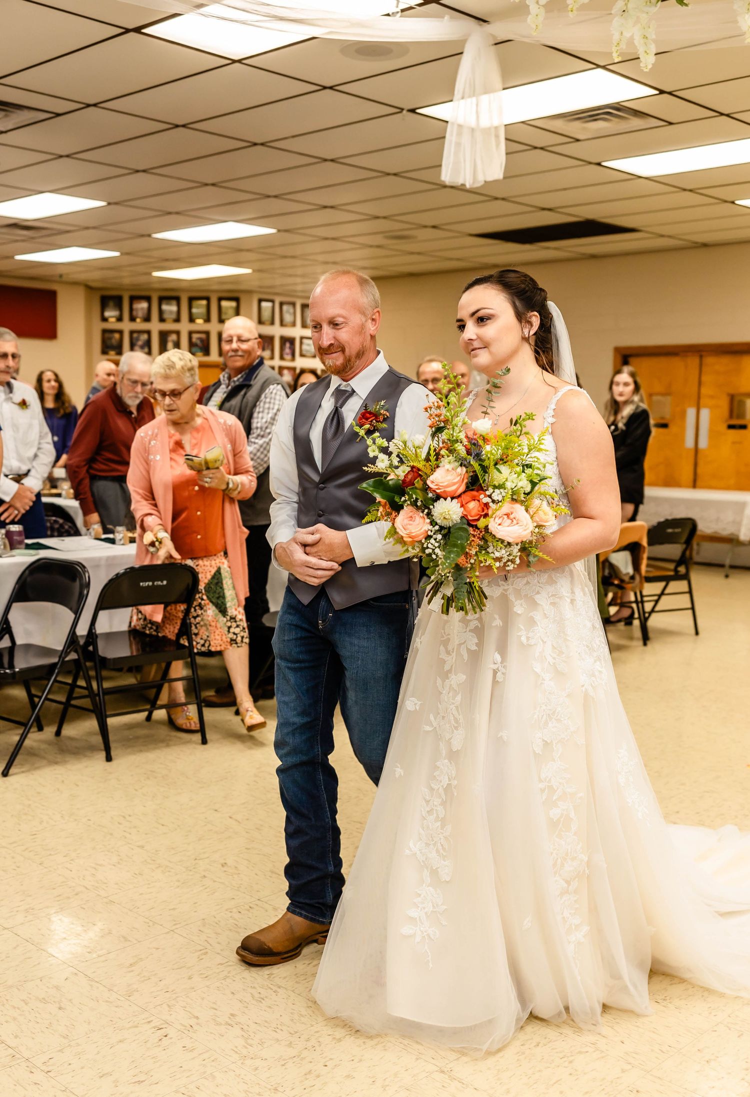A couple walks down the aisle during a wedding ceremony in a community hall with folding chairs.