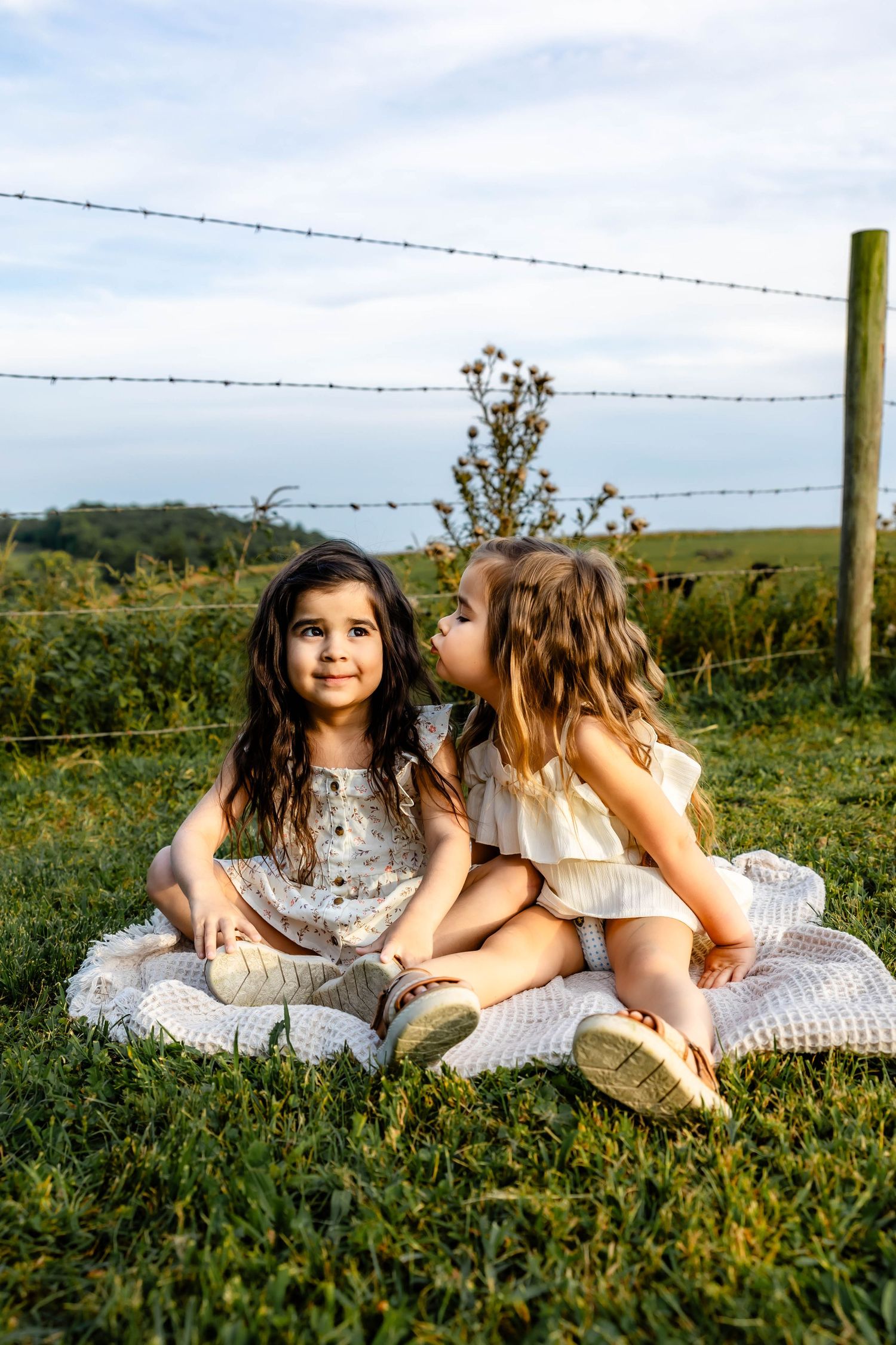 Two children in light colored dresses sitting together on blanket in pastoral setting during sunset.