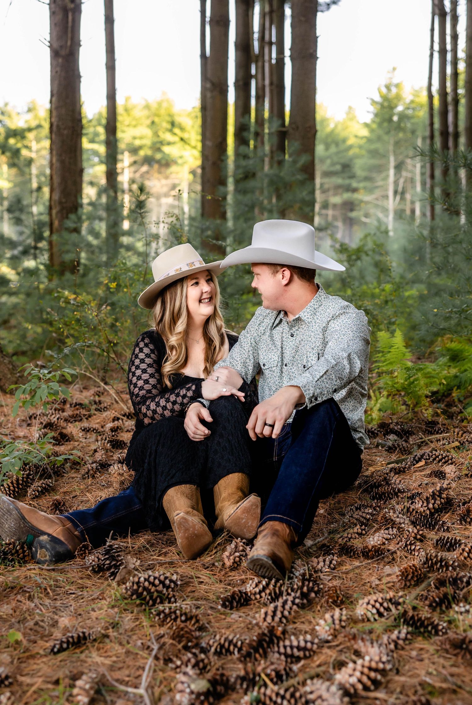 Couple wearing cowboy hats share sweet moment while sitting on forest floor covered in pine cones.