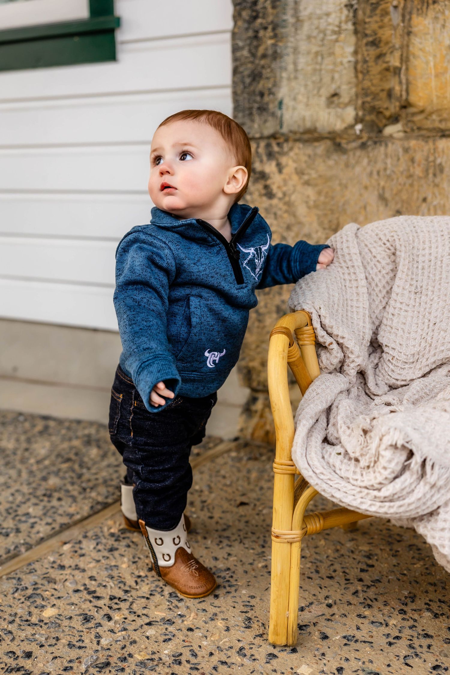 Small child in blue hoodie standing beside chair with knit blanket.