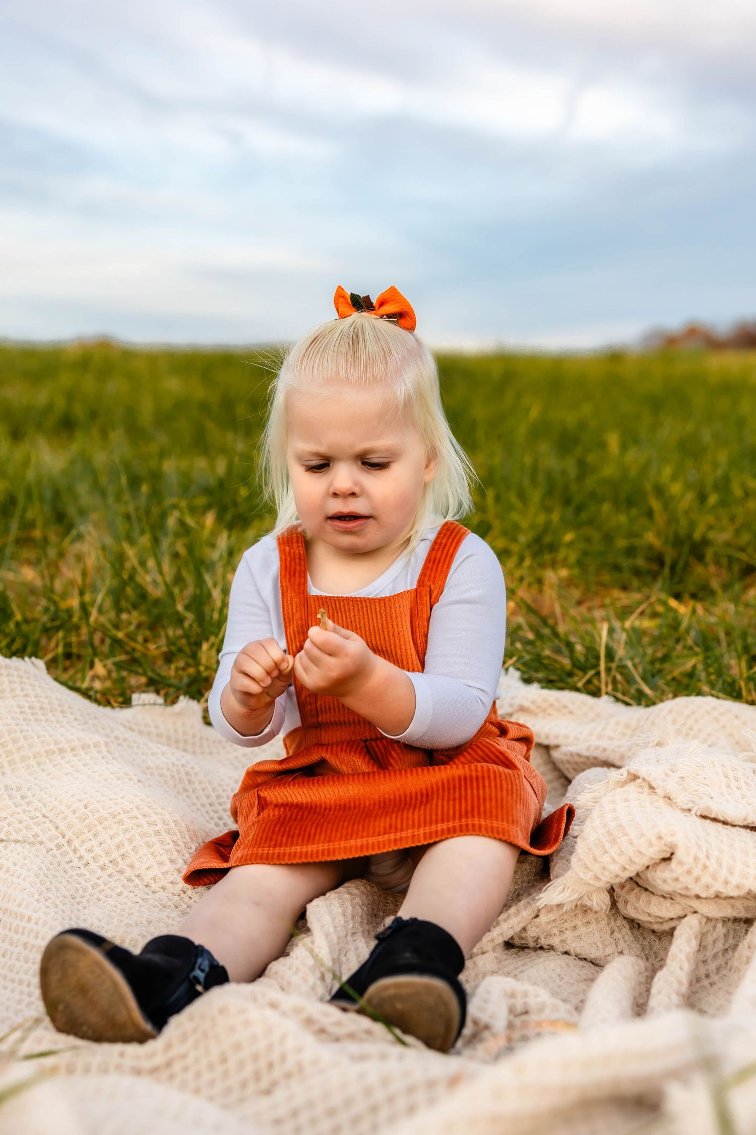 Child in orange corduroy dress sitting on cream blanket in grassy field.