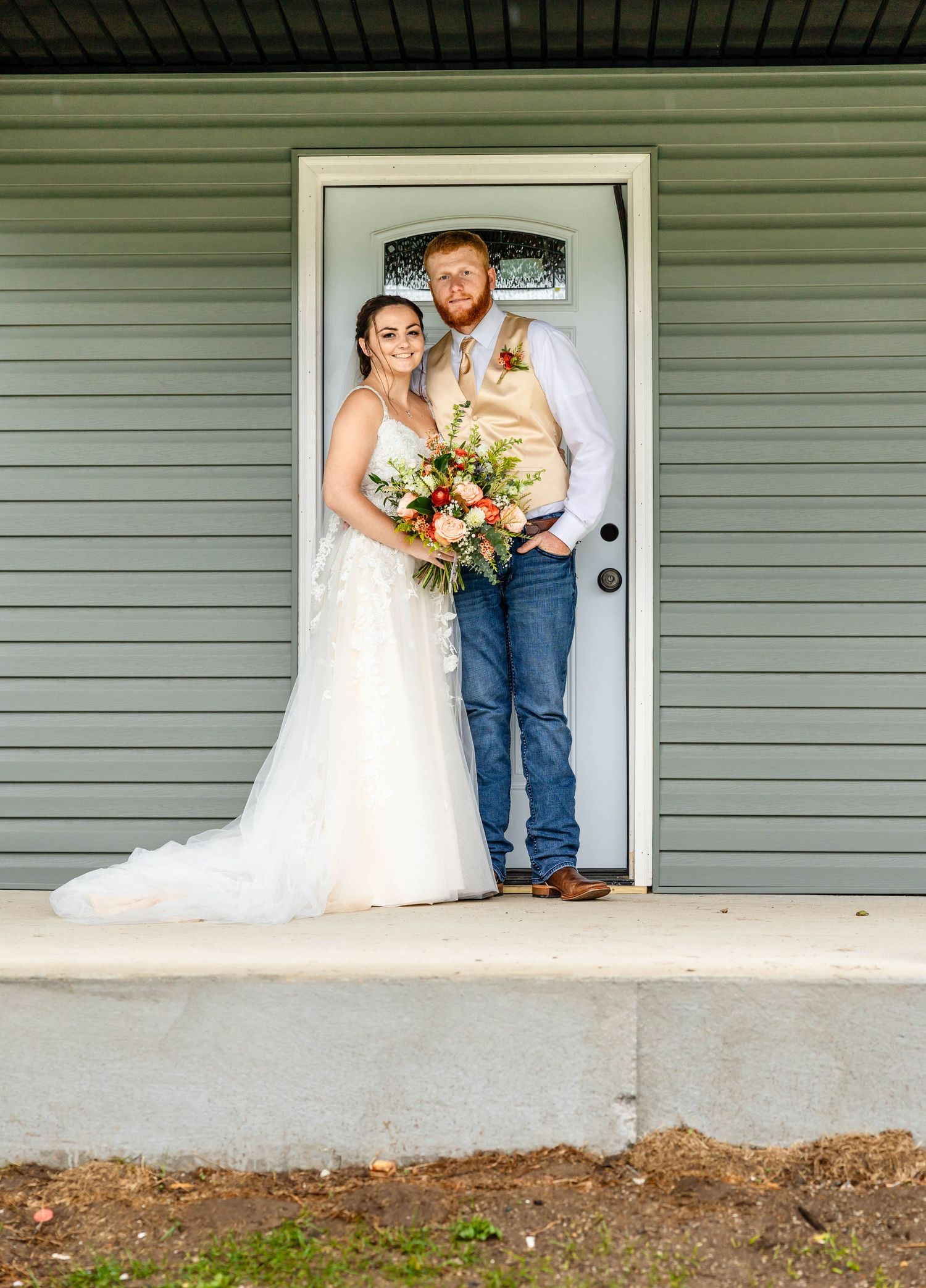 A couple poses in front of a sage green house holding a vibrant fall bouquet.