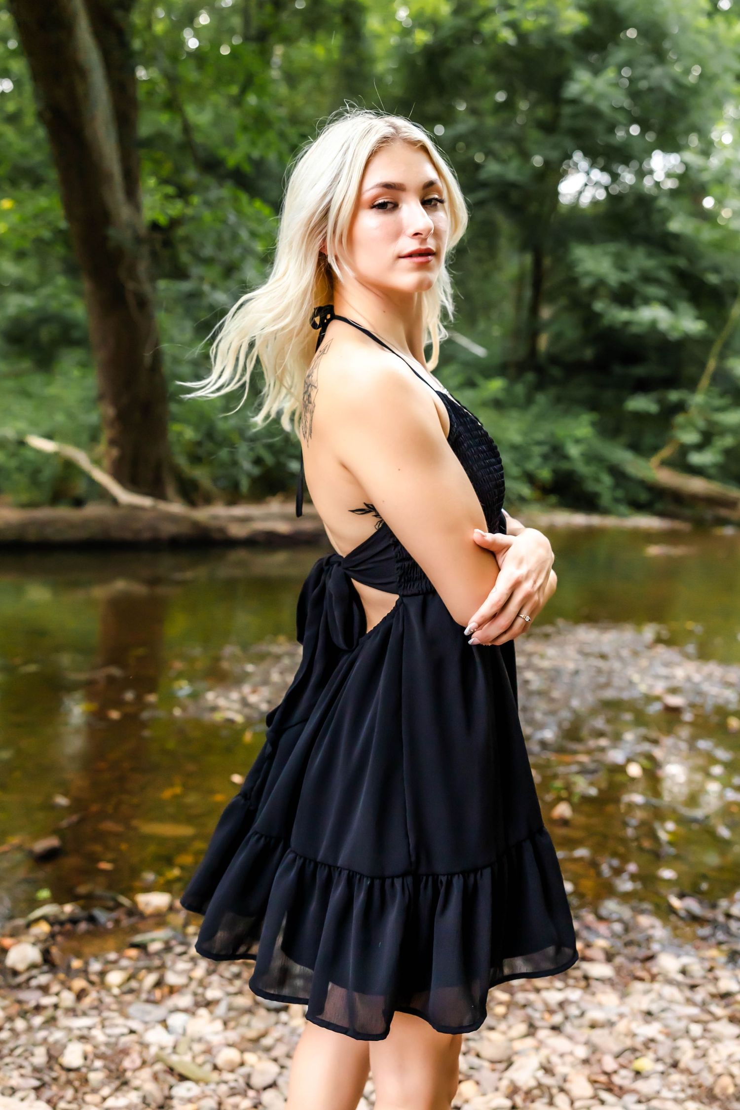 A black summer dress with side cutouts is displayed by a scenic creek with trees.