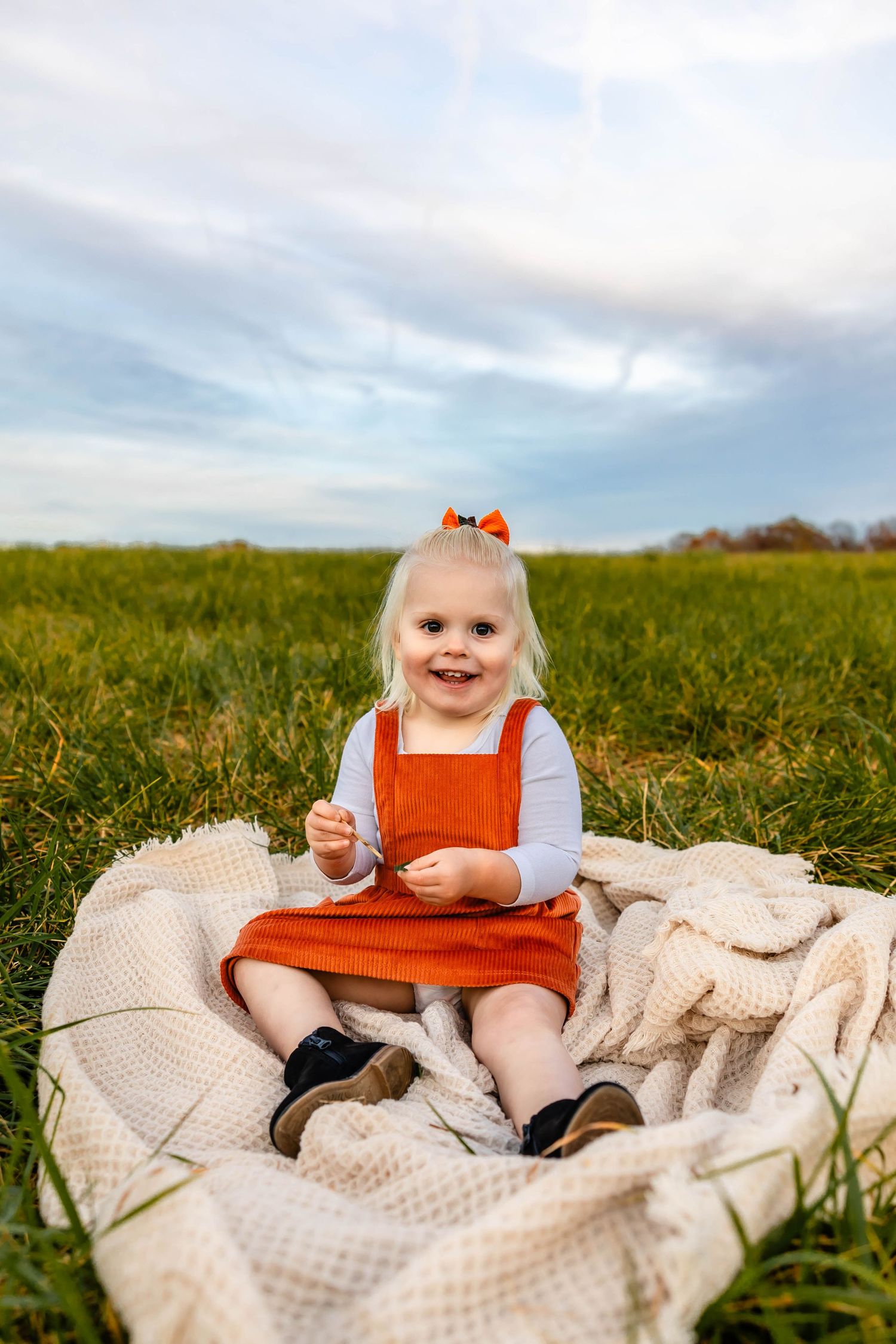 A cheerful toddler in orange overalls relaxes on a white blanket in a sunny meadow.