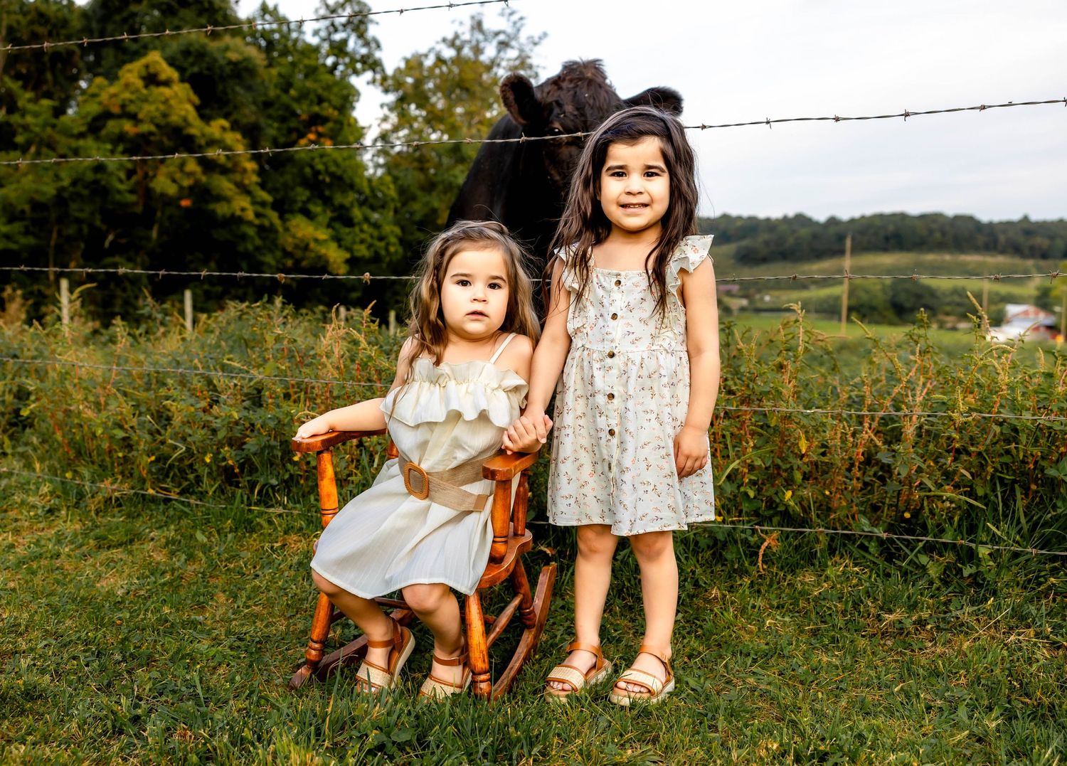Two children in summer dresses pose near barbed wire fence with cattle visible behind.