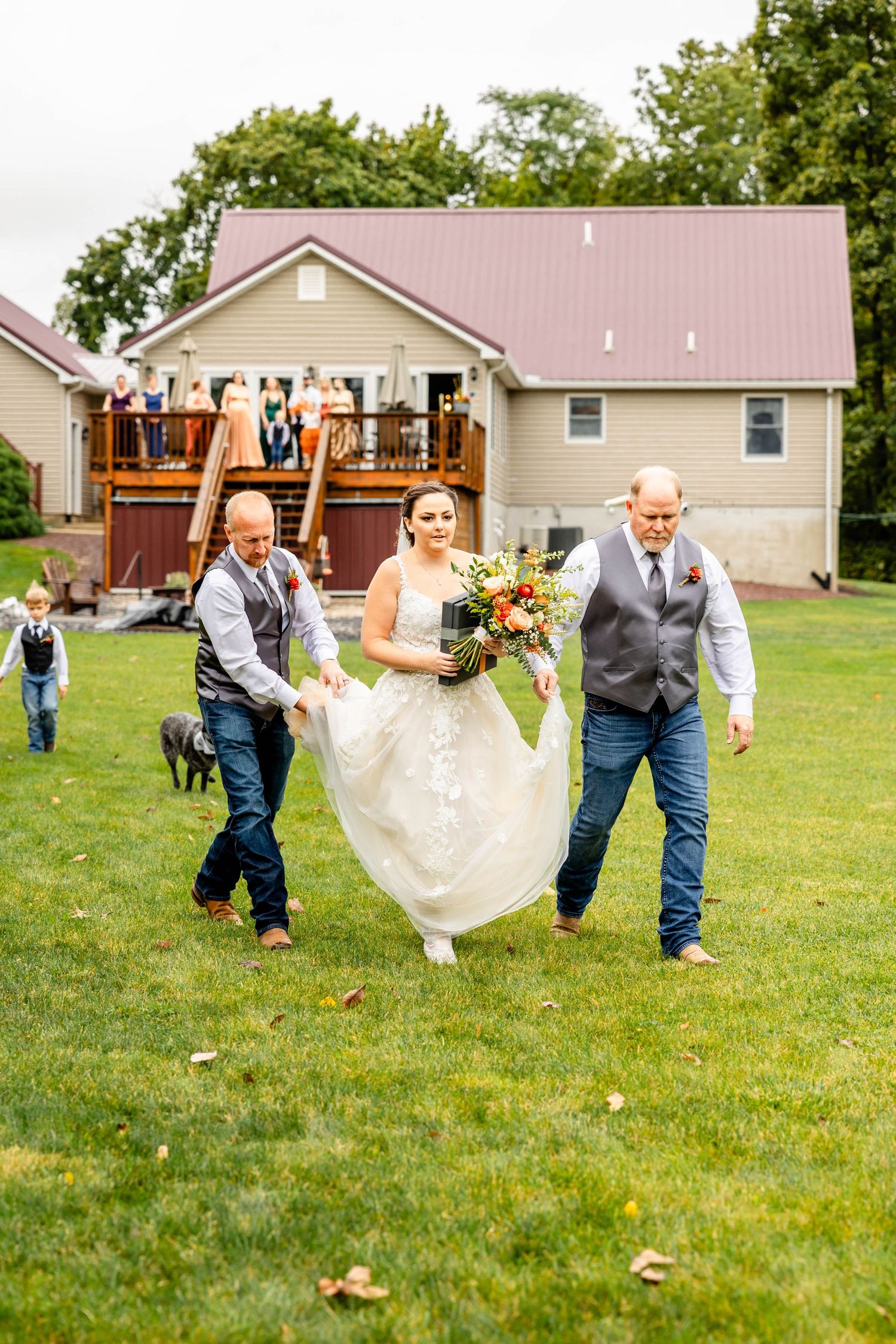 Wedding party helping carry the dress across a green lawn with a house in the background.