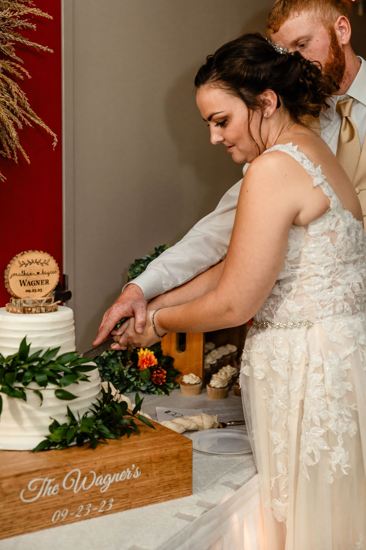 The wedding couple cuts their white wedding cake decorated with greenery and flowers.