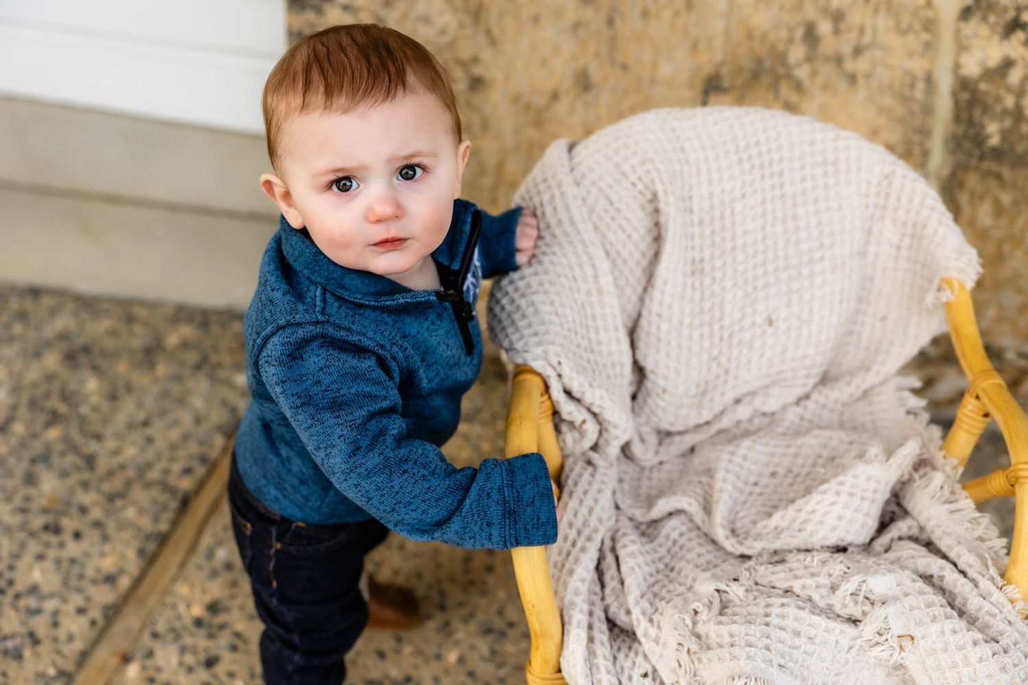 Young child in blue hoodie standing next to wooden chair with beige blanket.