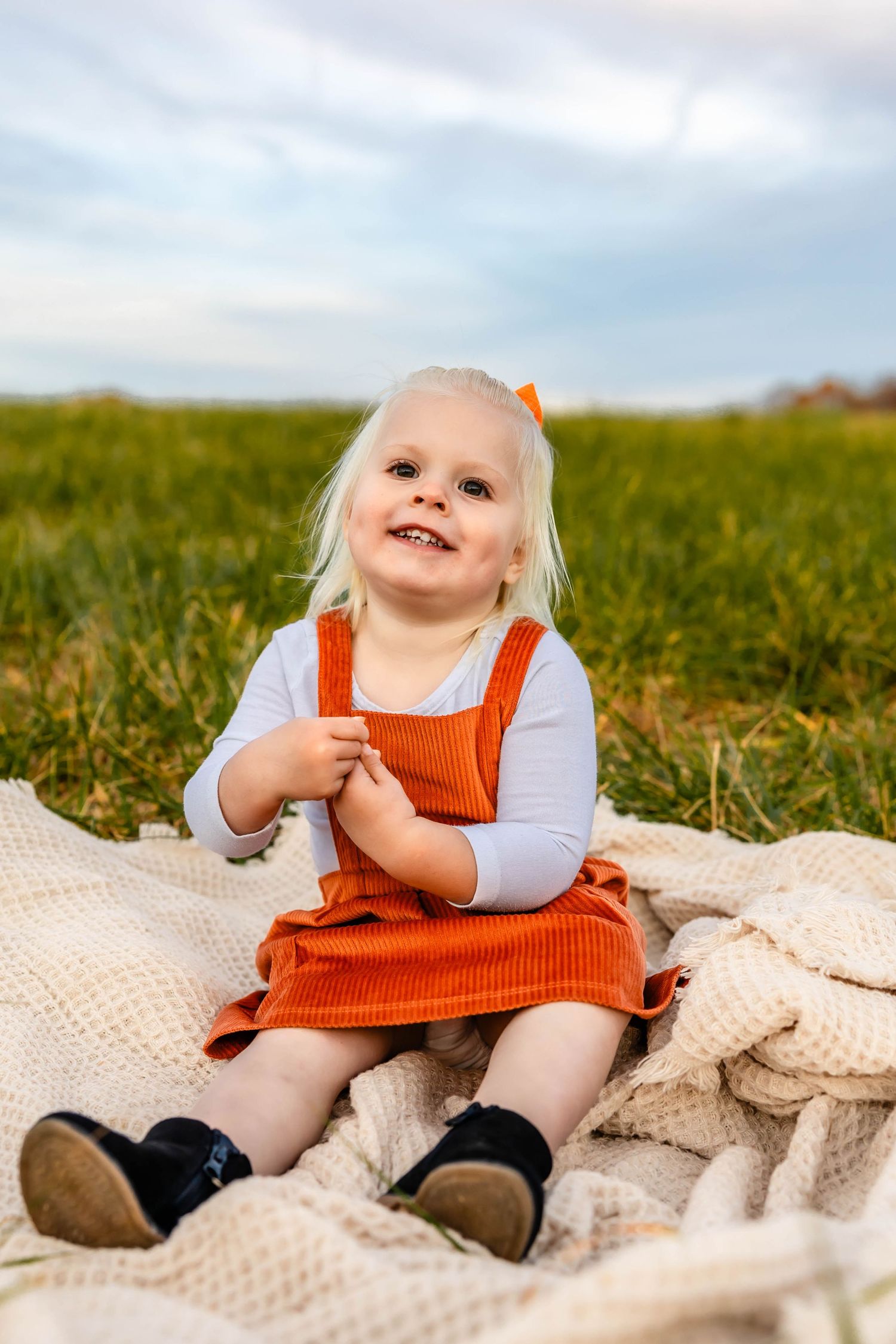 Toddler in orange jumper dress smiling while sitting on blanket in field.