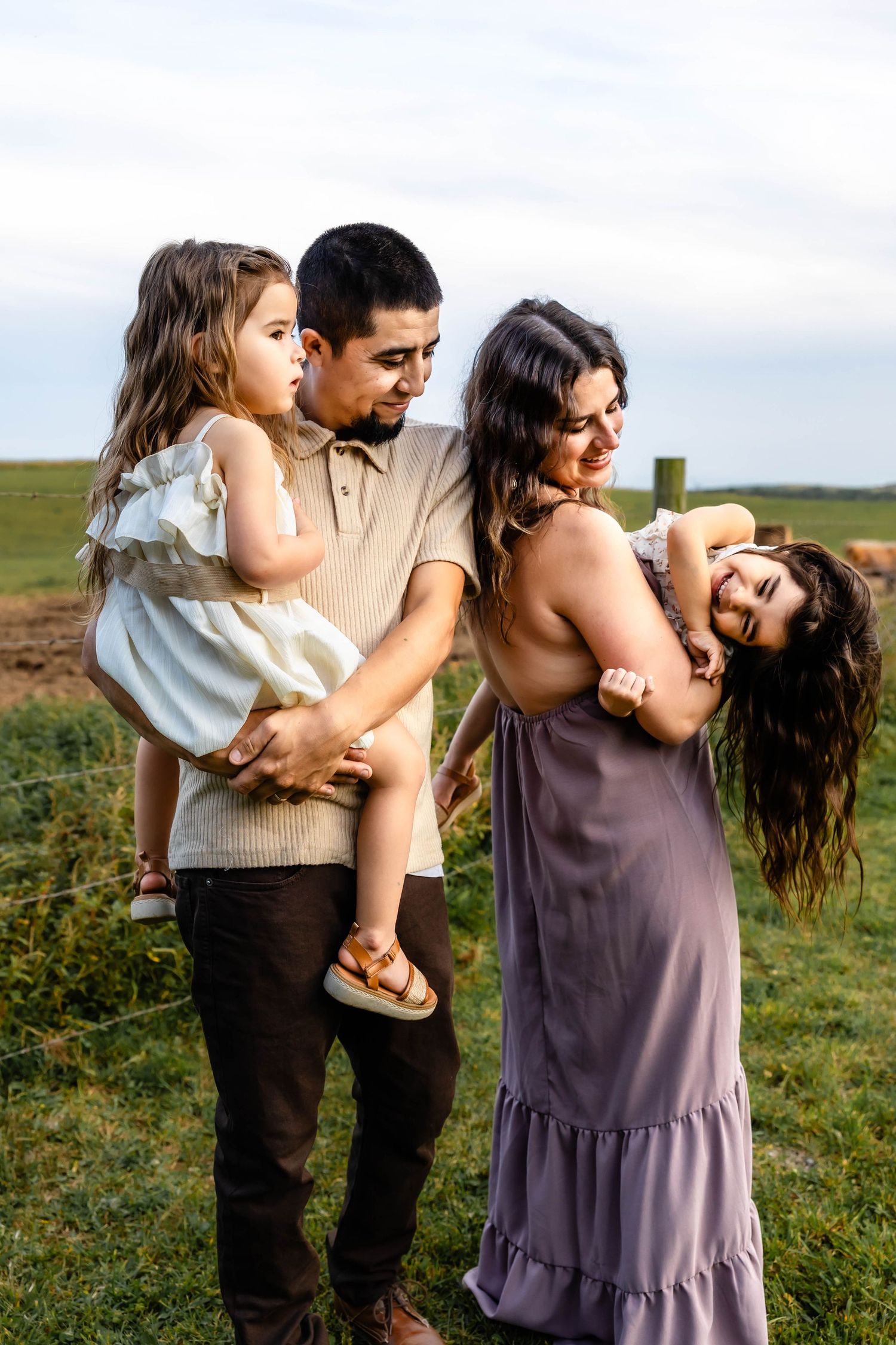 A playful family portrait outdoors with casual attire and natural lighting against a pastoral backdrop.
