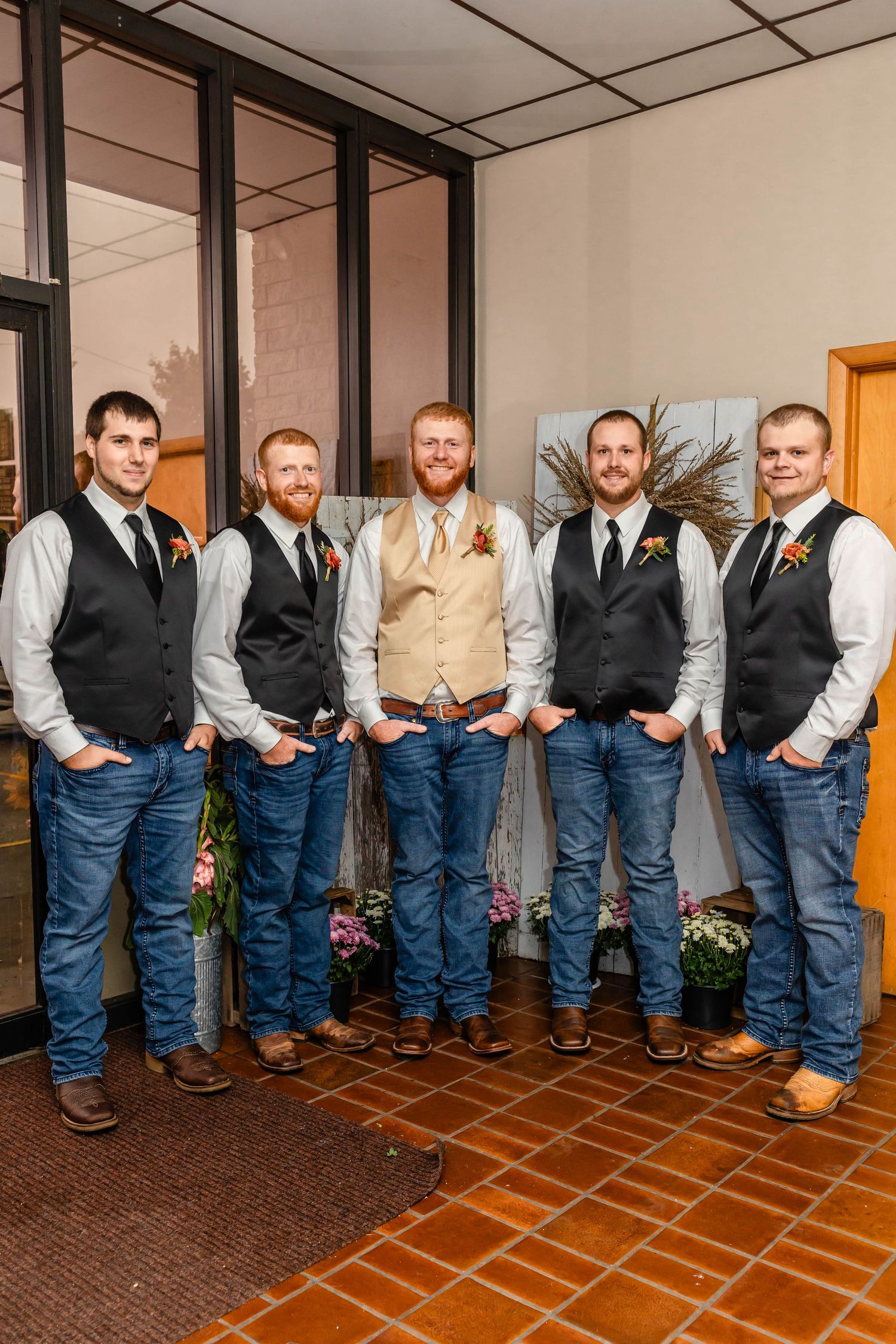 Groomsmen in vests and jeans pose together against indoor brick wall.