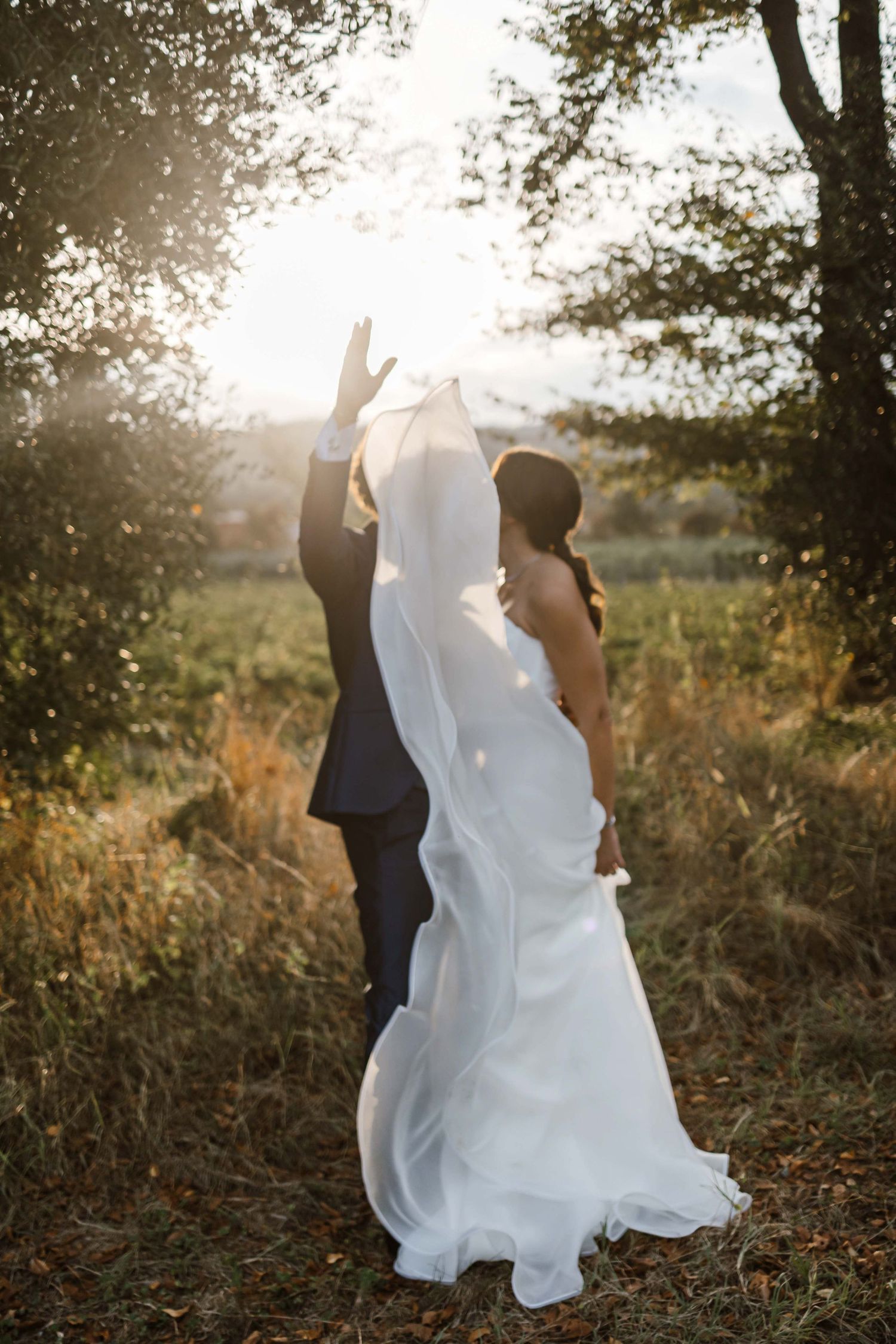 A romantic couple wearing wedding attire poses against golden sunlight in a natural outdoor setting.