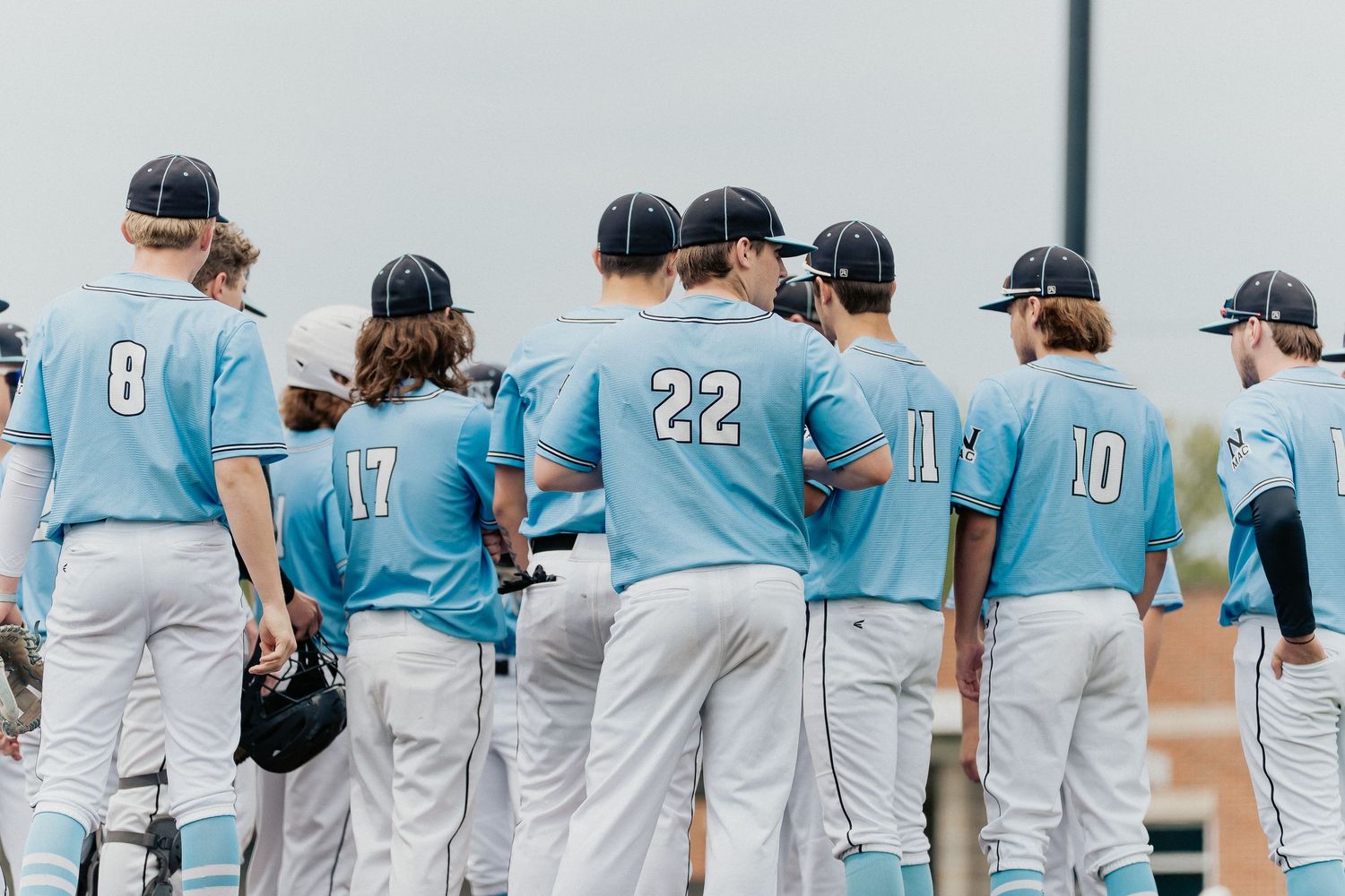 Baseball team players gather wearing light blue jerseys with numbers showing.