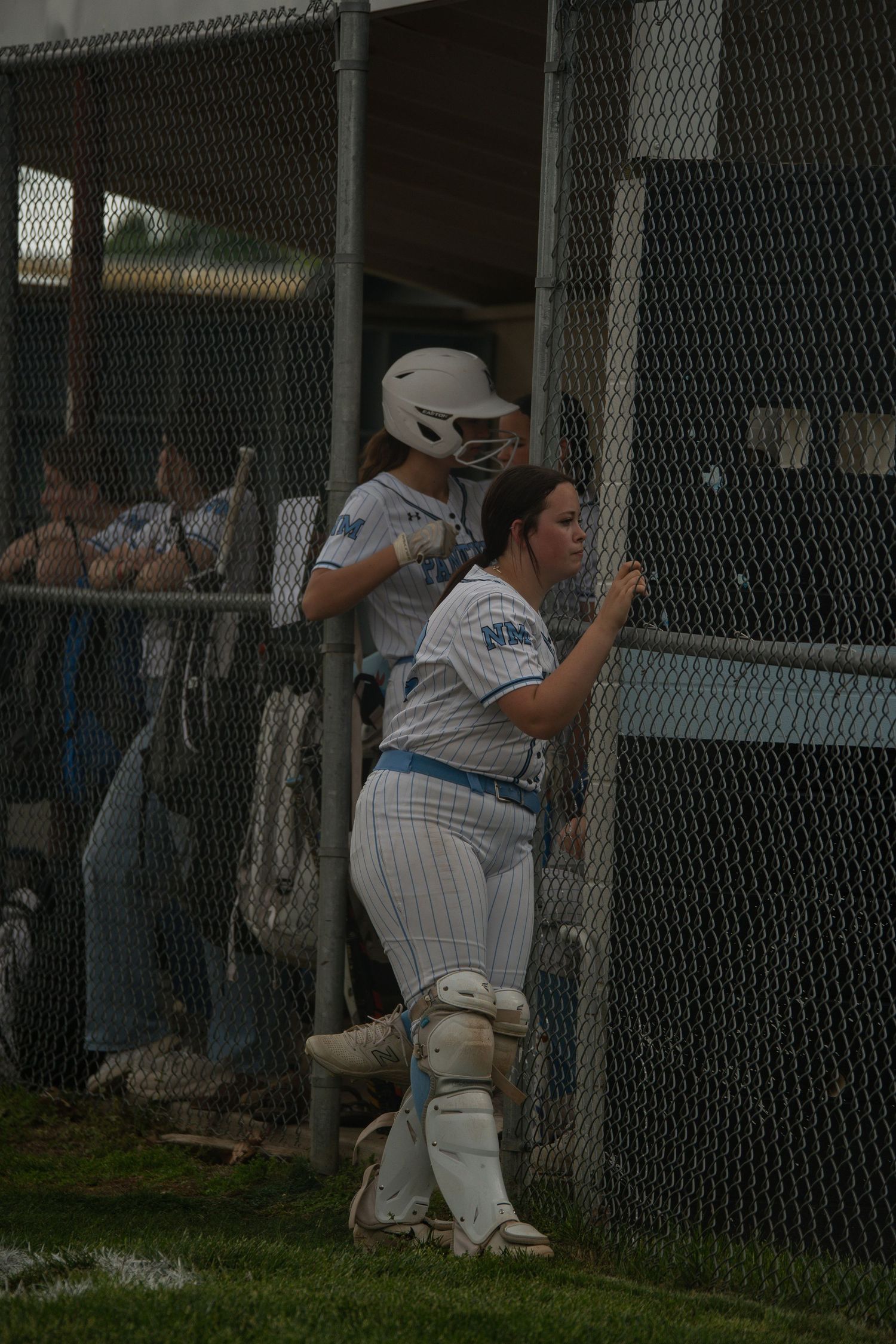 Baseball players watching the game through dugout fence in evening light.