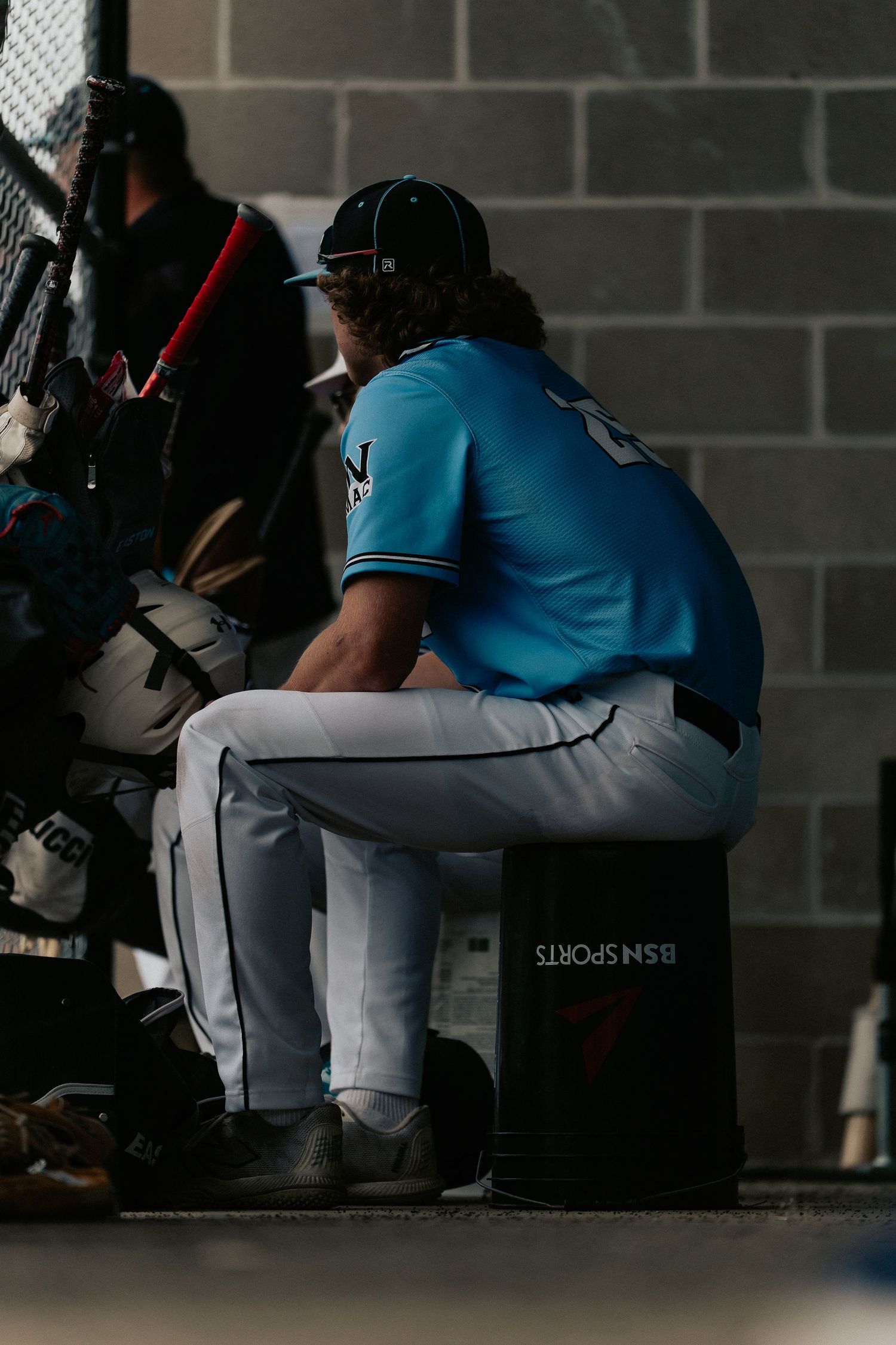 Baseball player in light blue uniform sits on bucket in dugout during game.