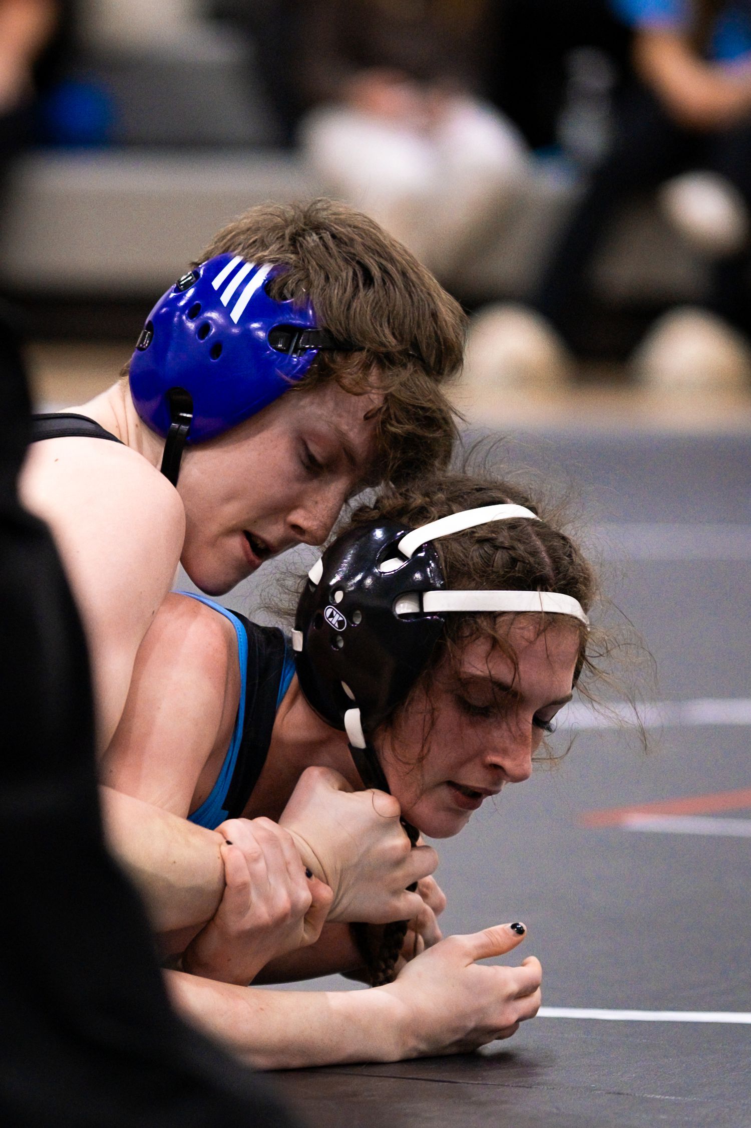Two wrestlers in blue and black headgear grapple on the mat during a match.
