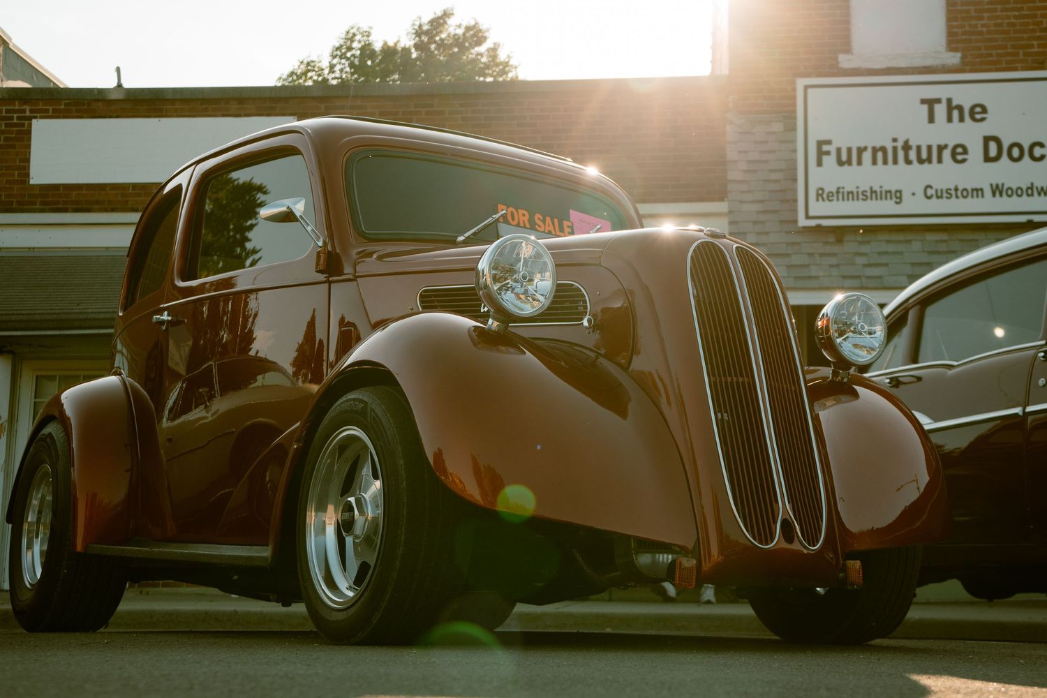 Vintage brown hot rod car parked in front of The Furniture Doc store at sunset.