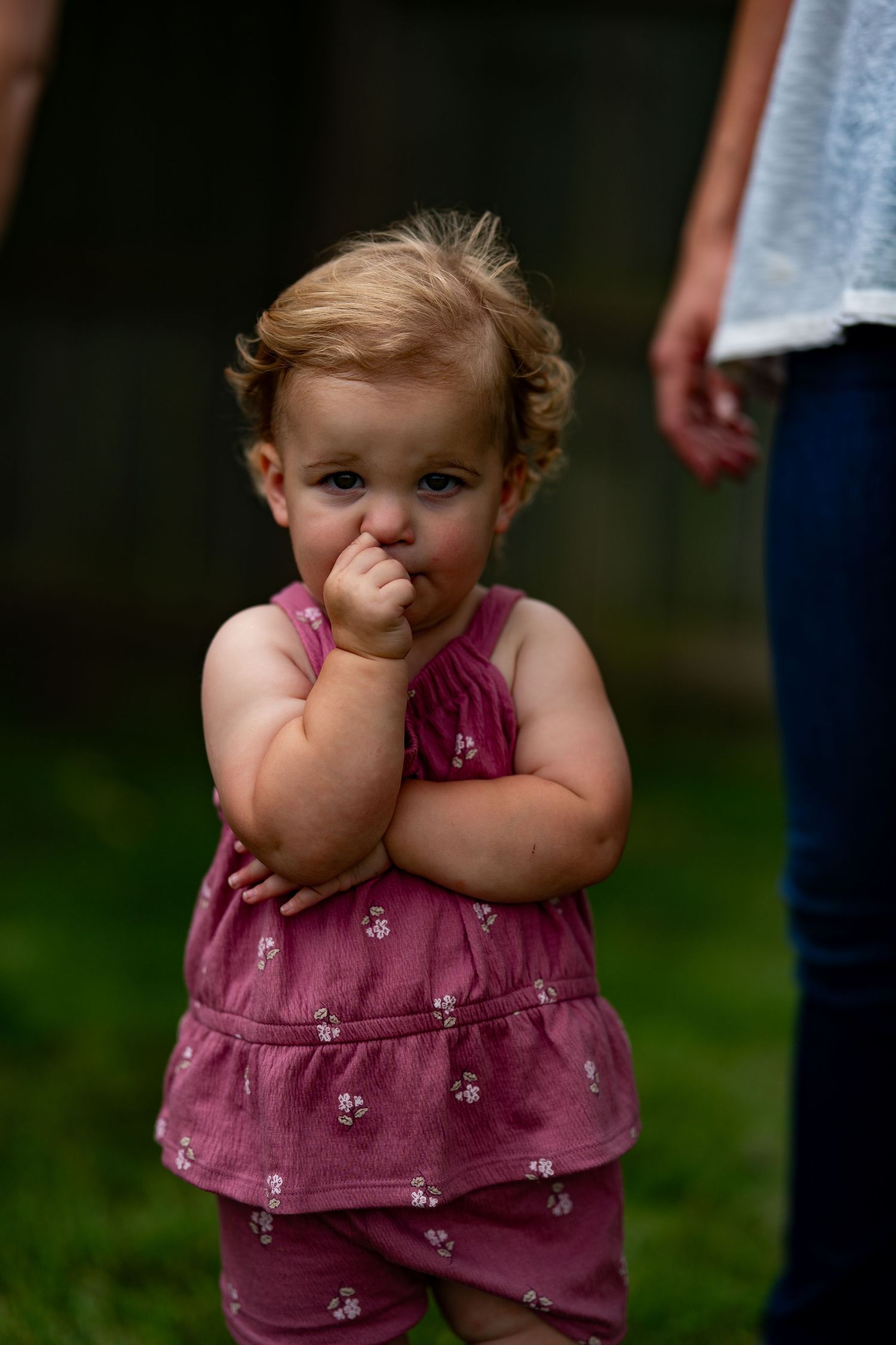A small child in a pink floral dress standing outdoors with a finger near their mouth.