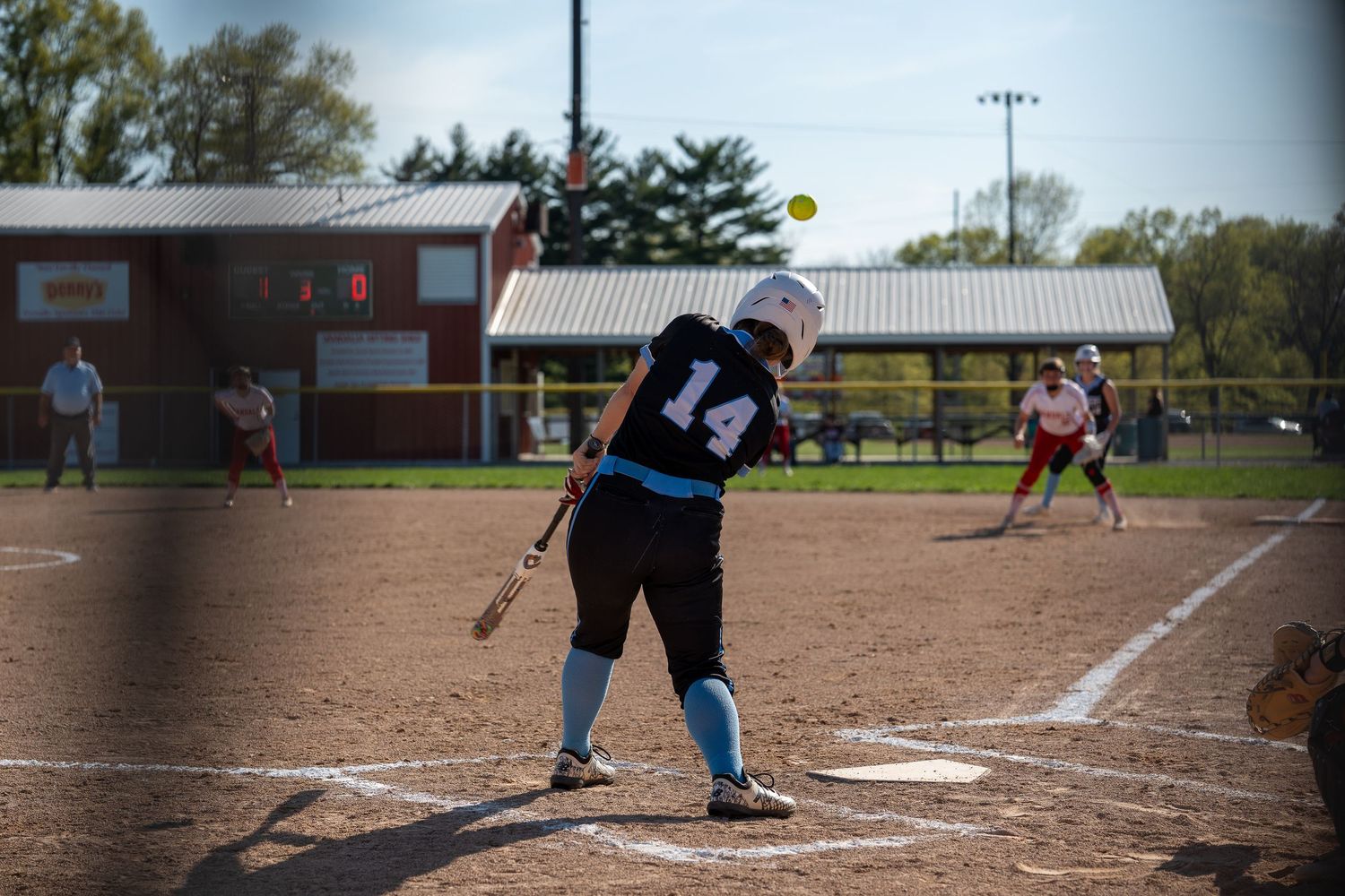 Softball player at bat on a dirt field during a game at dusk.
