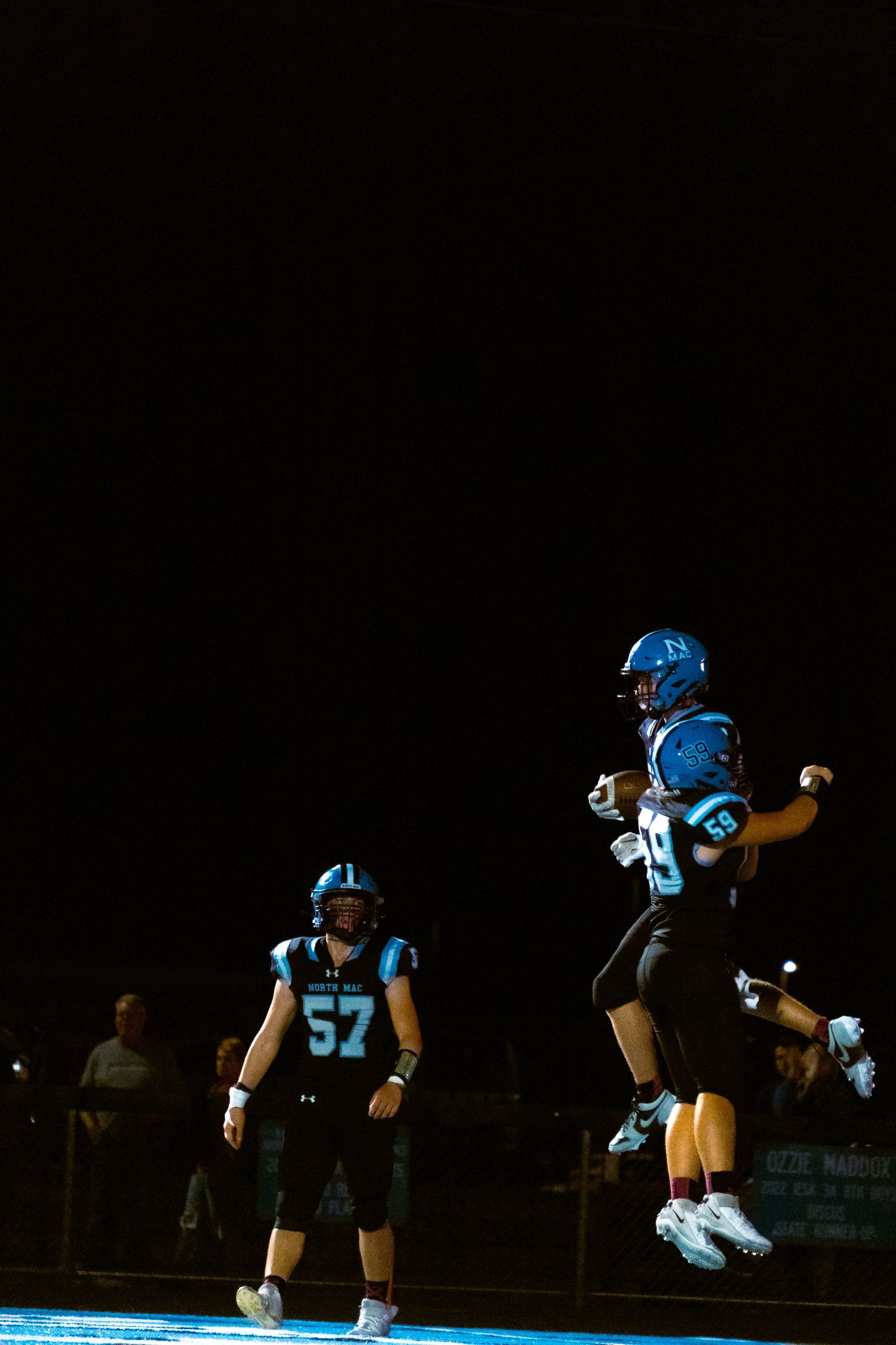 Football players in bright blue uniforms on a dark field with one athlete jumping high in celebration.