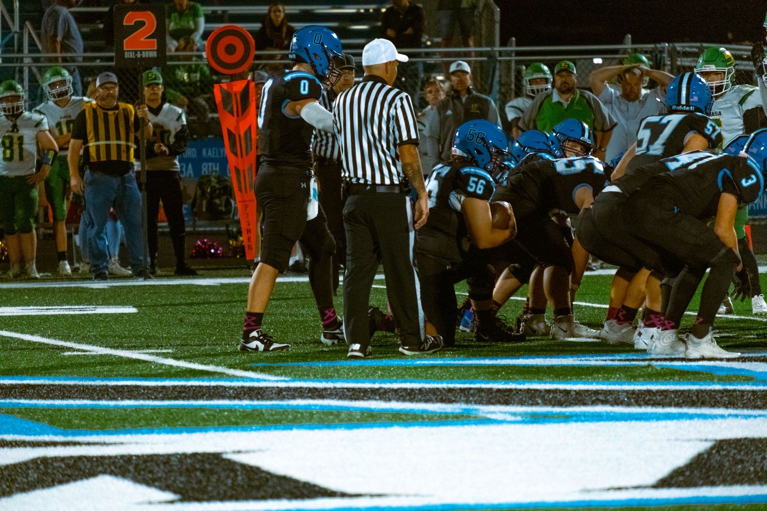 Football players in orange and blue uniforms gather around an official on the field during a night game.