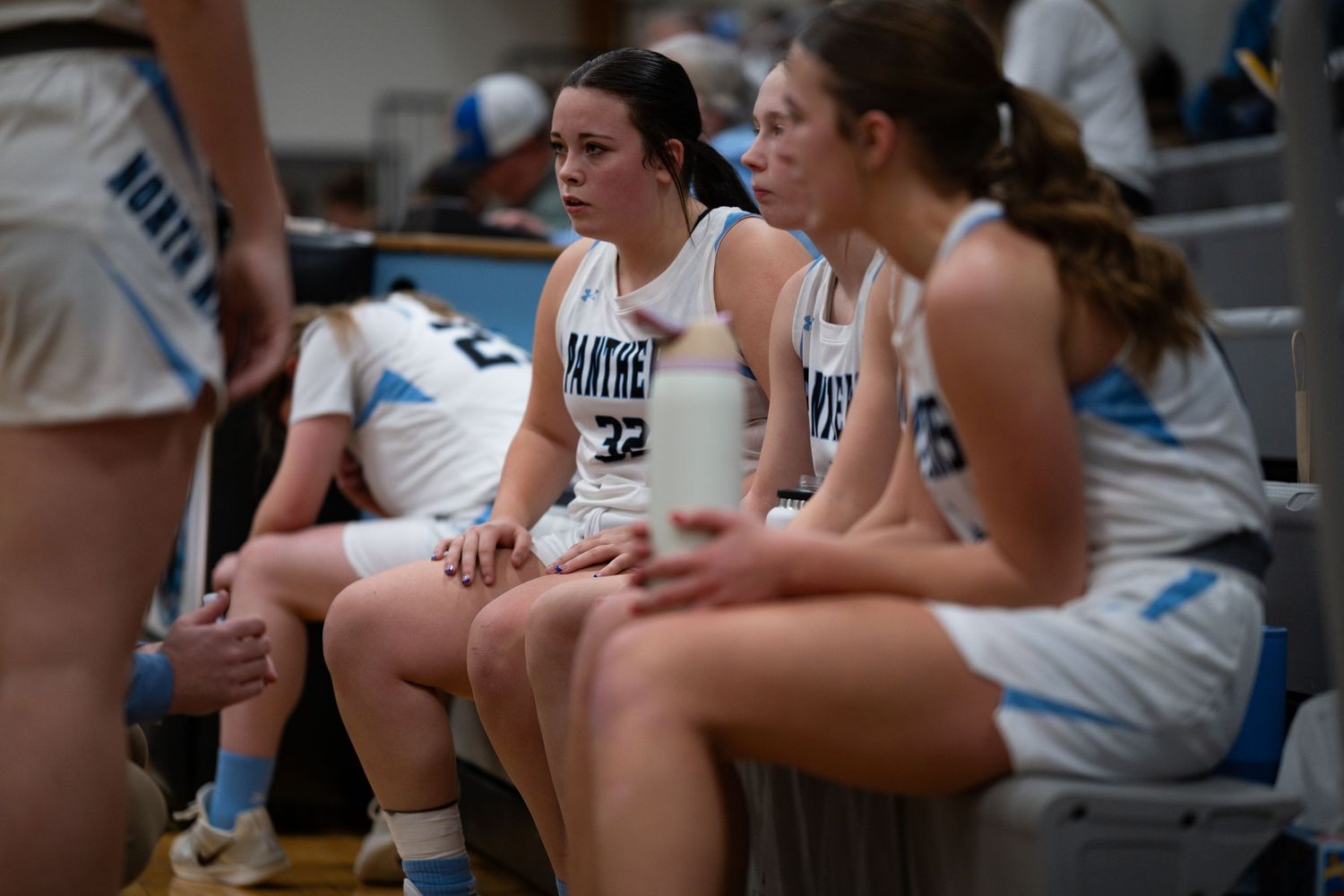 Basketball players sit together on the sideline bench during a game wearing light colored uniforms.