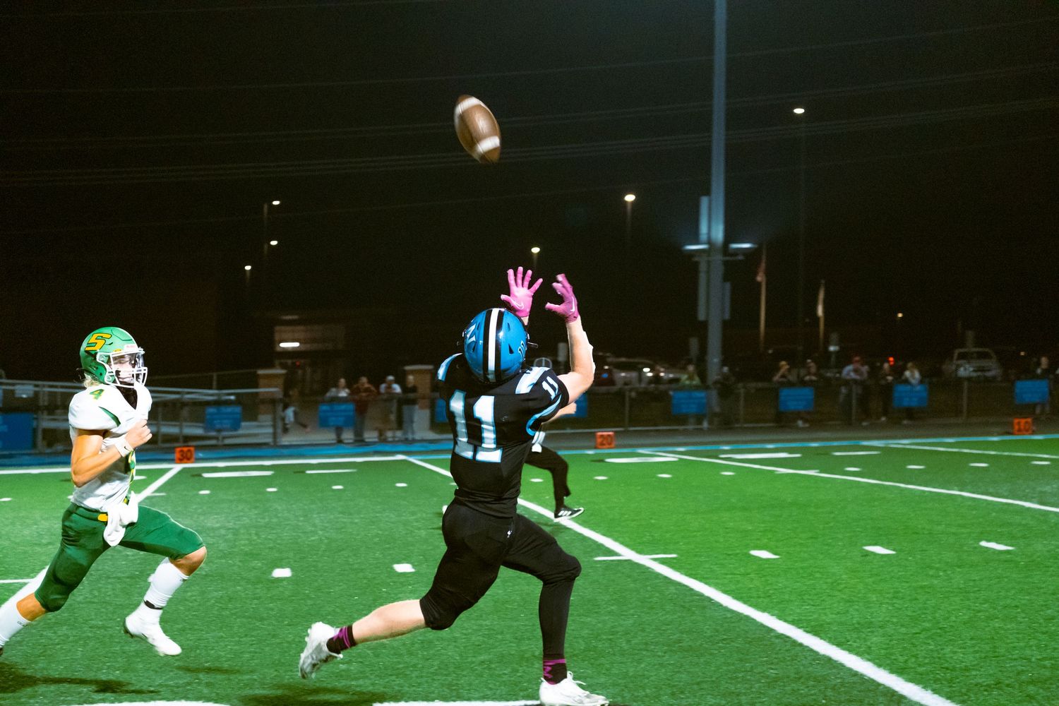 Football player in black uniform catches a pass while being pursued by defender in white uniform during night game.