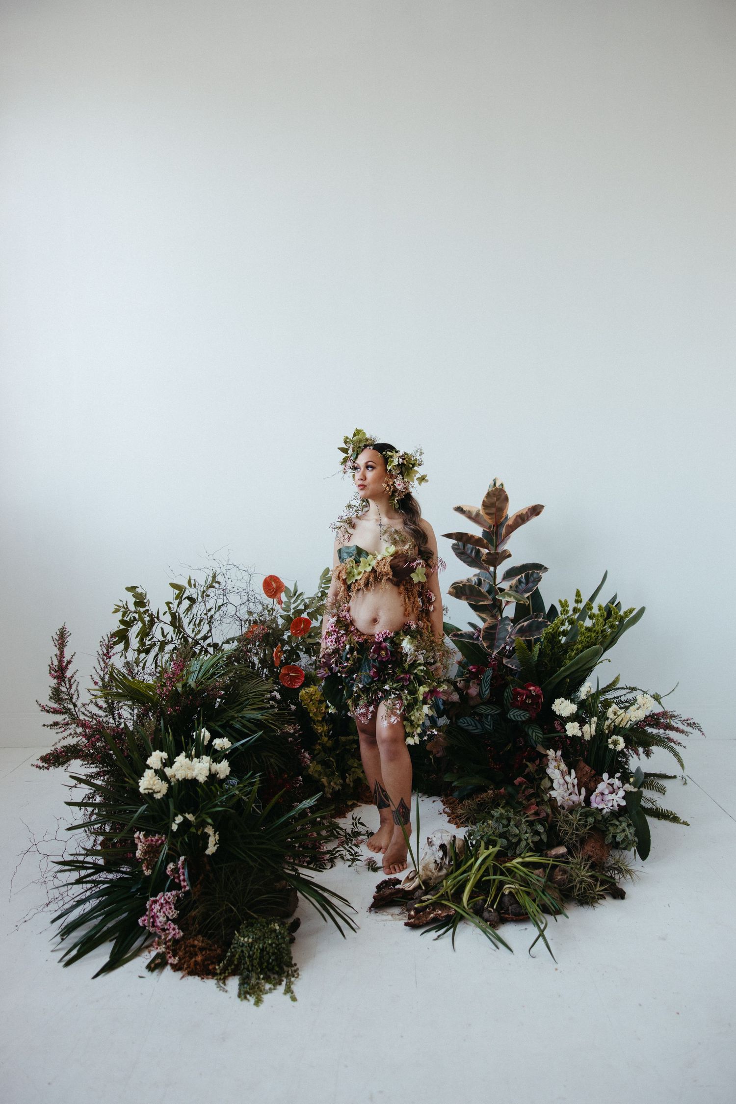Artistic studio portrait with model standing on lush floral display against white backdrop adorned with flowers and greenery.