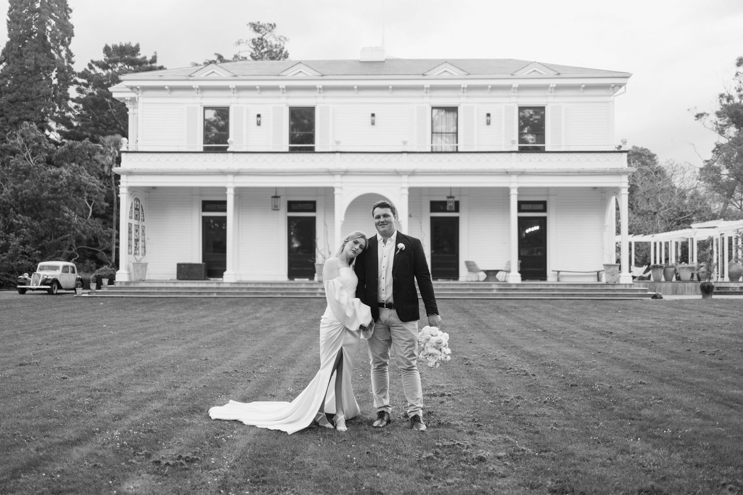 Wedding couple poses with dog on lawn in front of historic white colonial building.
